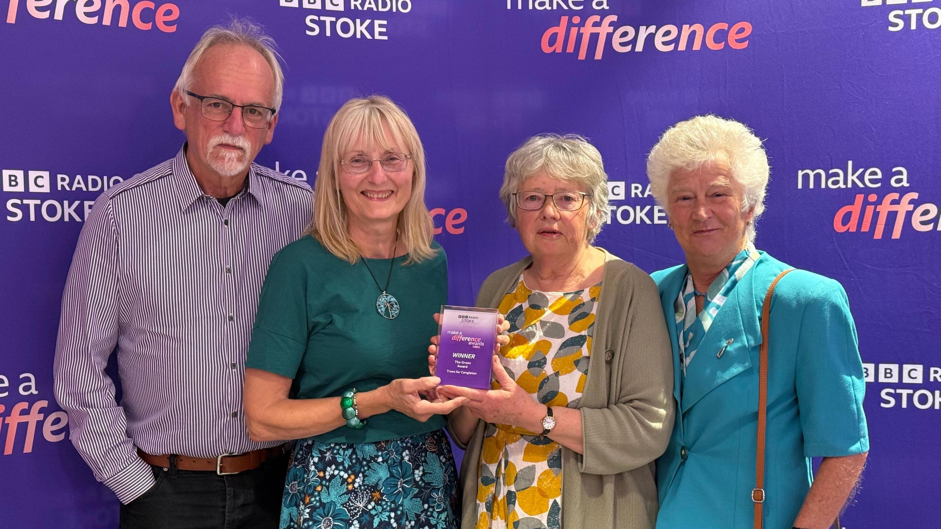 A man and four women are stood in a row, with the two women in the middle holding a glass award. They are standing in front of a purple BBC Radio Stoke Make A Difference step and repeat board.