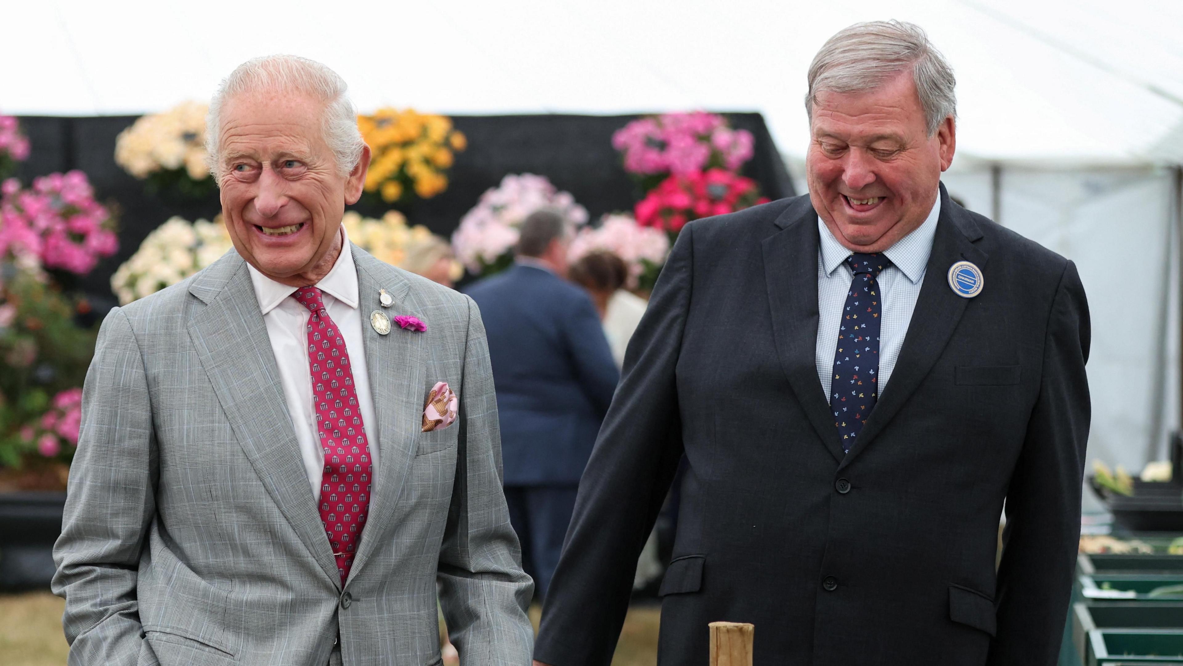 King Charles II walking and laughing with Graham Browne, the chairman of the Sandringham Flower Show. King Charles III is wearing a pale grey suit with a burgundy coloured tie. Mr Browne is wearing a black suit with a navy tie. Behind them is a wall of flowers. 