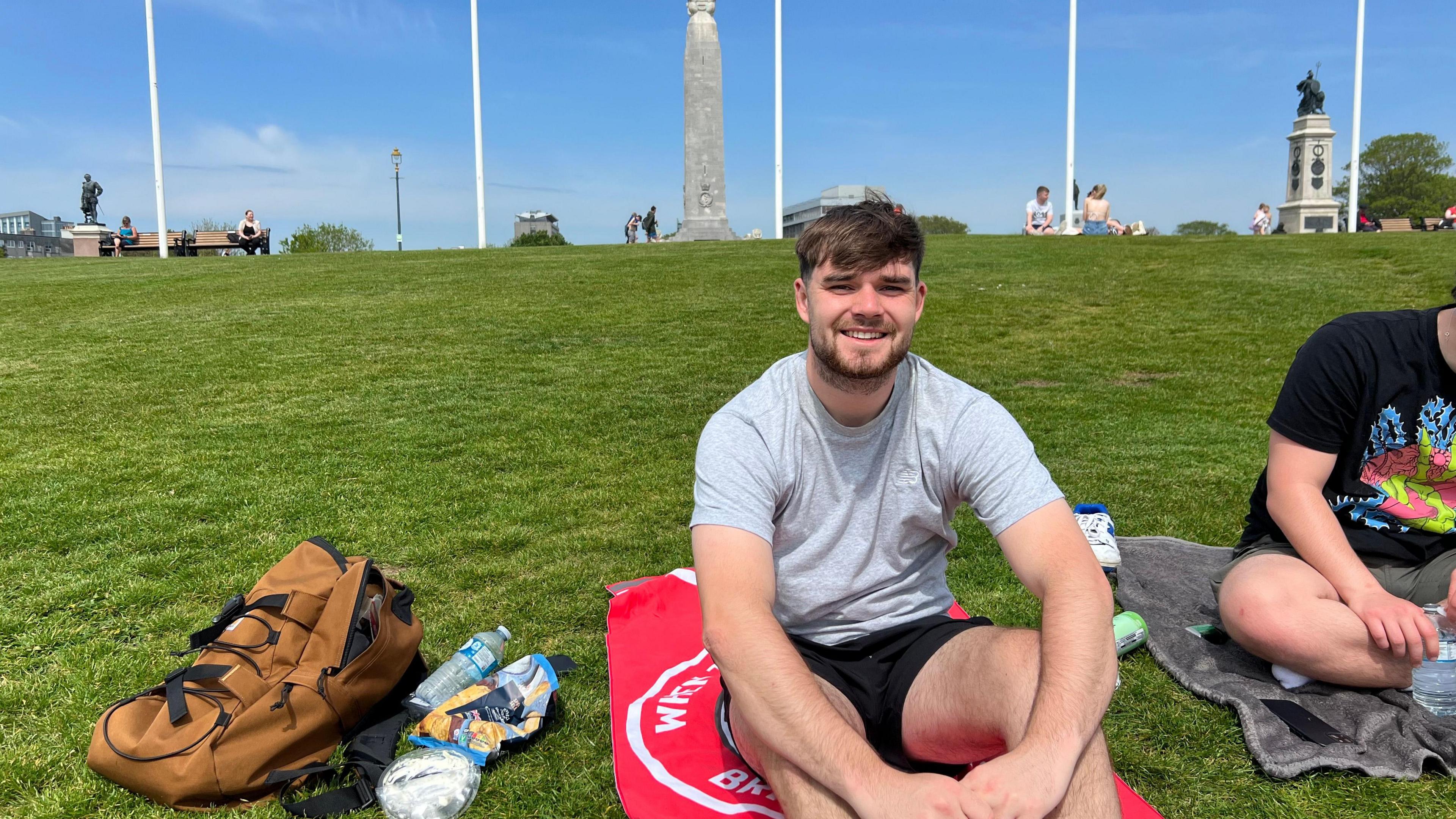 Jude Harris sat on a red towel, on the grass, smiling at the camera. Beside him is a brown rucksack.