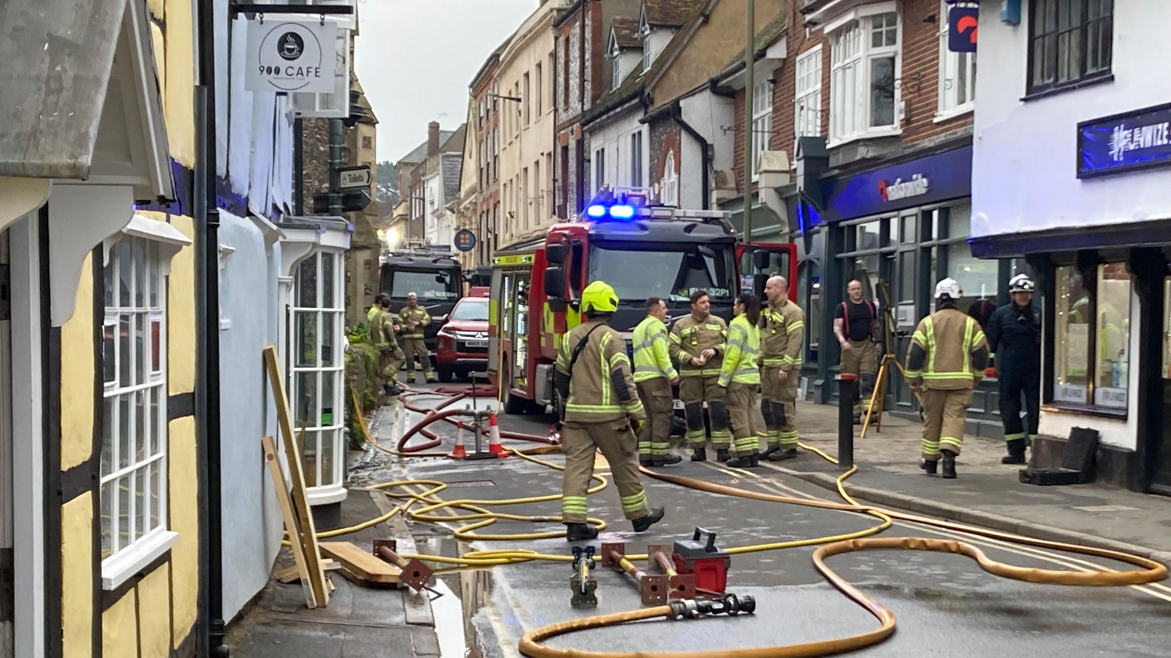 Around seven firefighters gather together on a small urban street. Several yellow hoses can be seen on the ground going from the engines into the front door of a small terraced building, which has the sign 900 Cafe on it. A fire engine is visible in the background. 