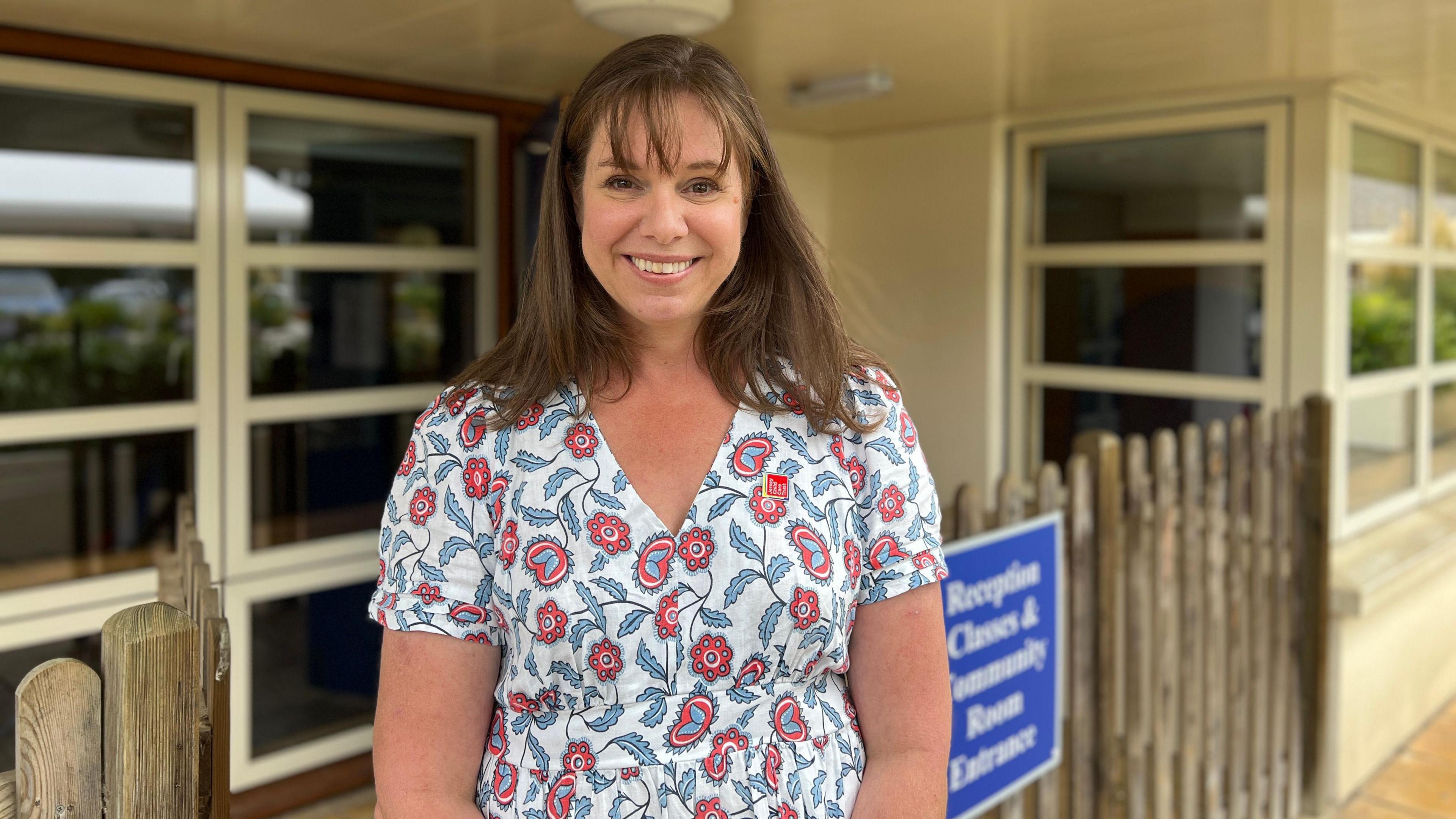 Fiona Vacher - a woman with brown hair and a fringe, smiling with teeth wearing a white t shirt with paisley pattern of blue and red, behind is a blurred background of a school building and a fence