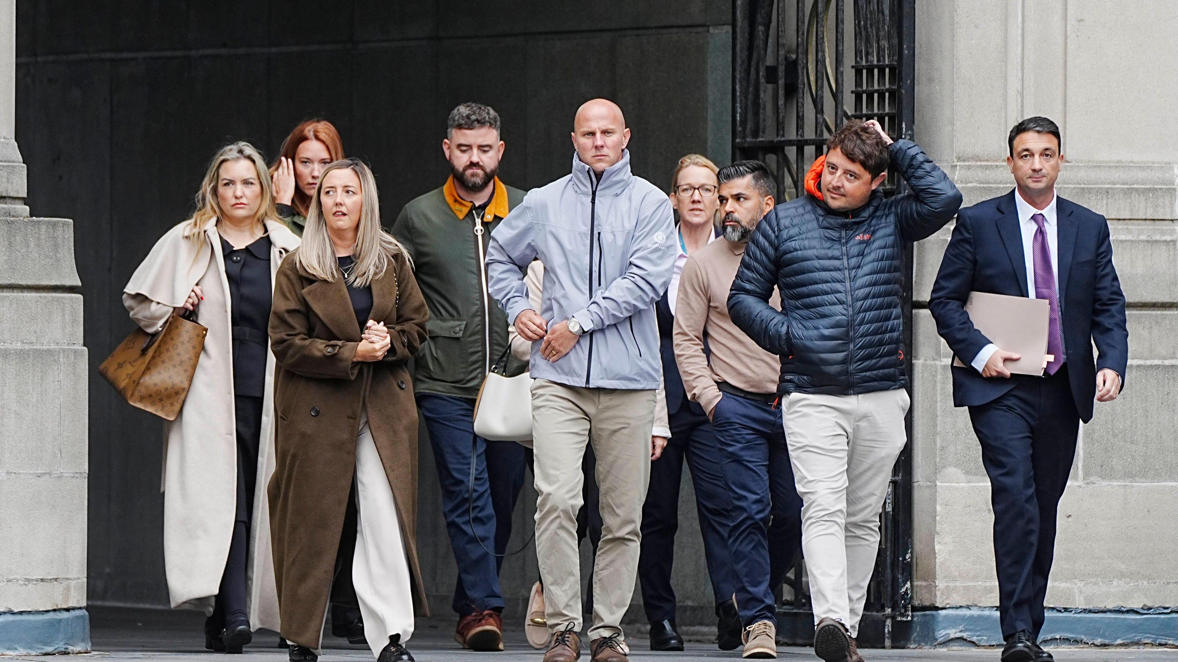 A group of nine people, including four women and five men, cross a stone square with sombre, determined expressions.