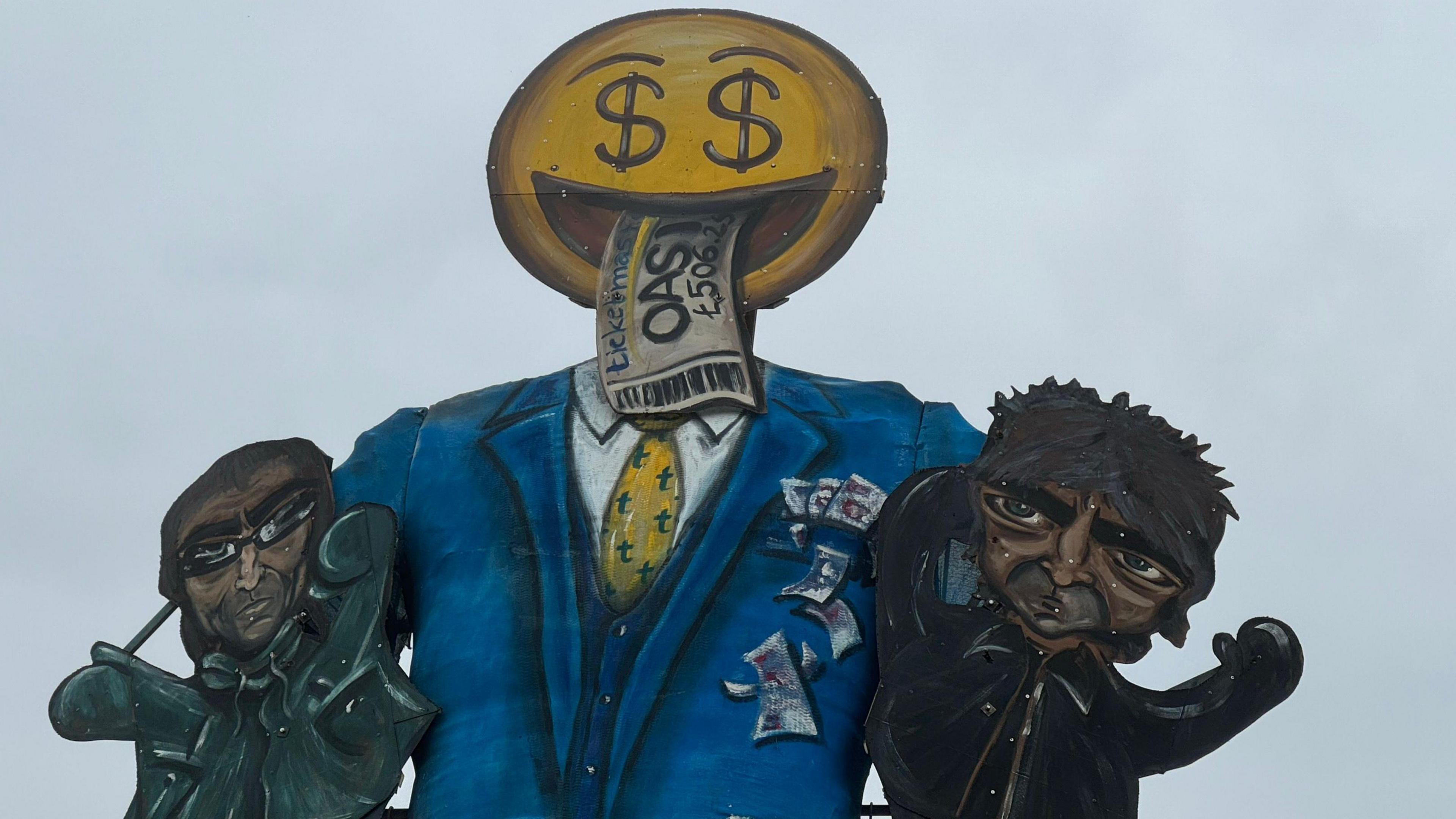 An effigy show Noel and Liam Gallagher as puppets to a man in a blue suit with a yellow smiley emoji head and dollar signs for eyes