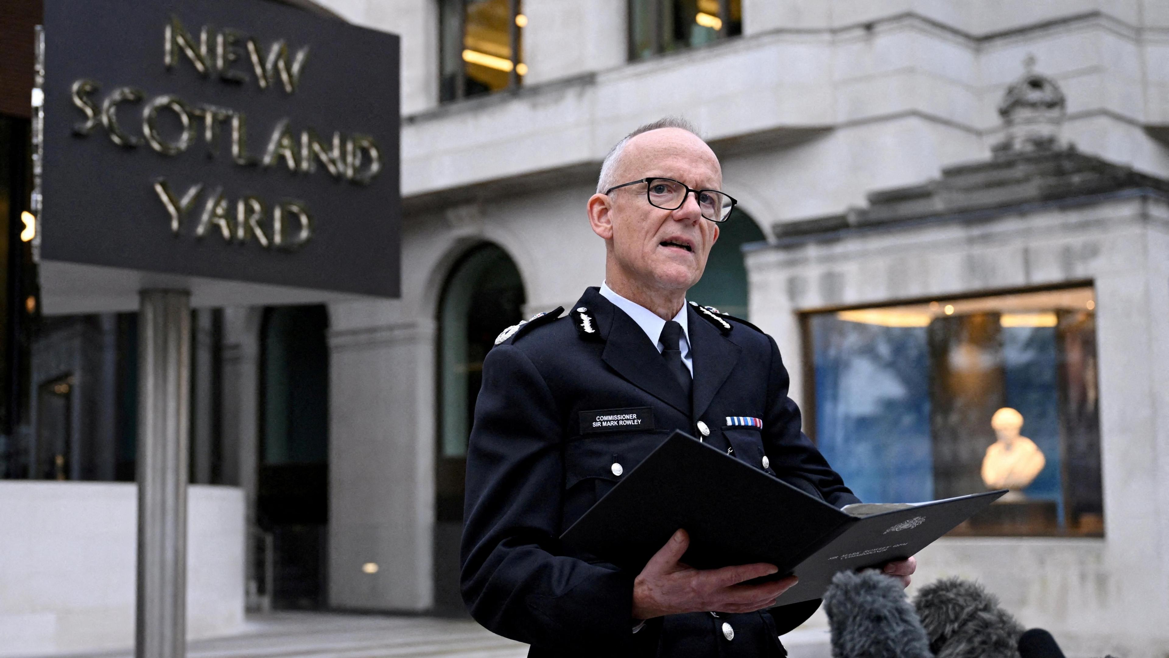 Sir Mark Rowley, a man with short grey hair and black glasses, stands in front of New Scotland Yard in his police uniform holding a black folder and speaking into microphones.