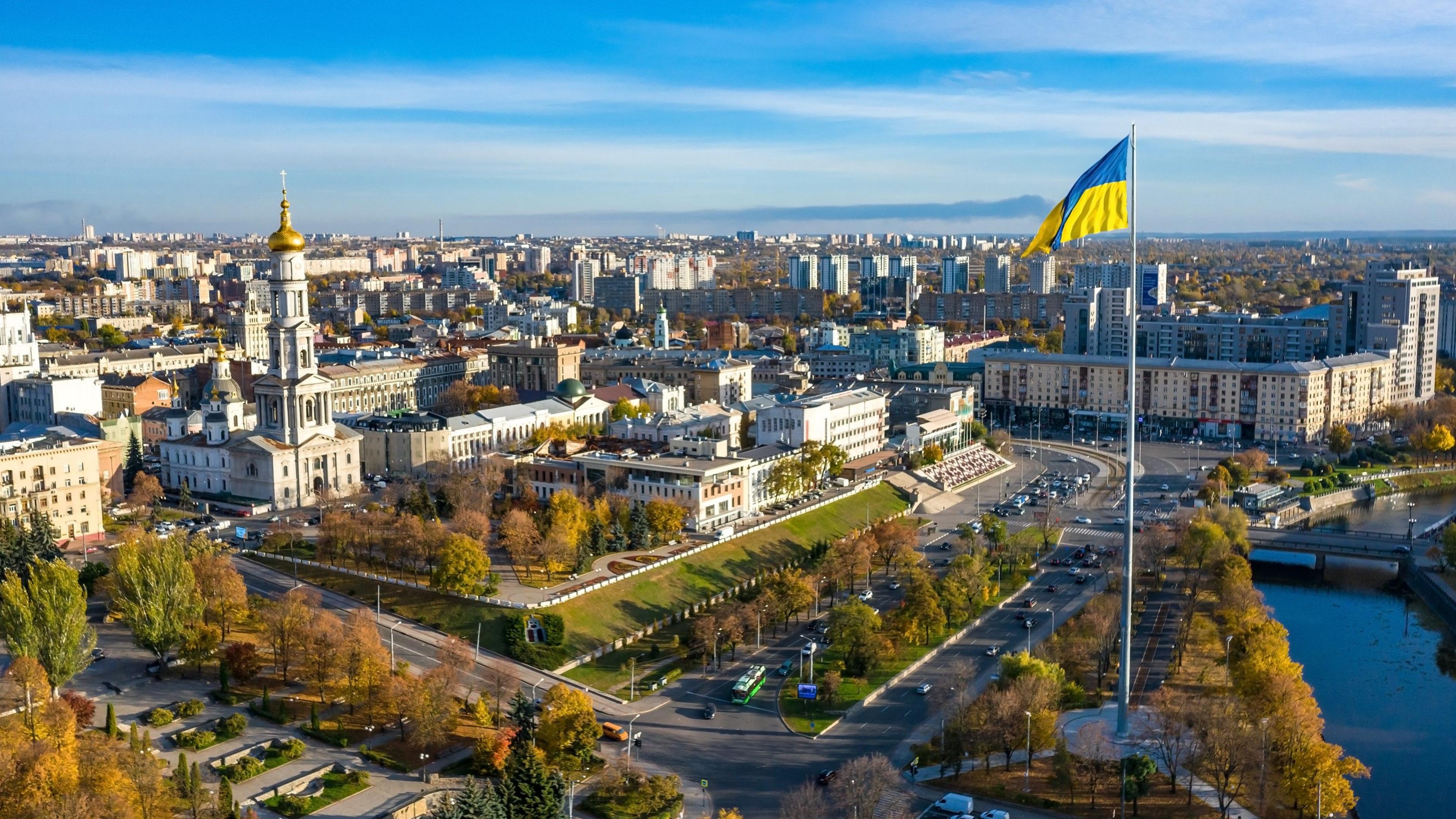 Aerial shot showing the Ukrainian flag flying above the city of Kharkiv, with wide avenues, churches and other buildings. There are trees, grassy areas and a river.