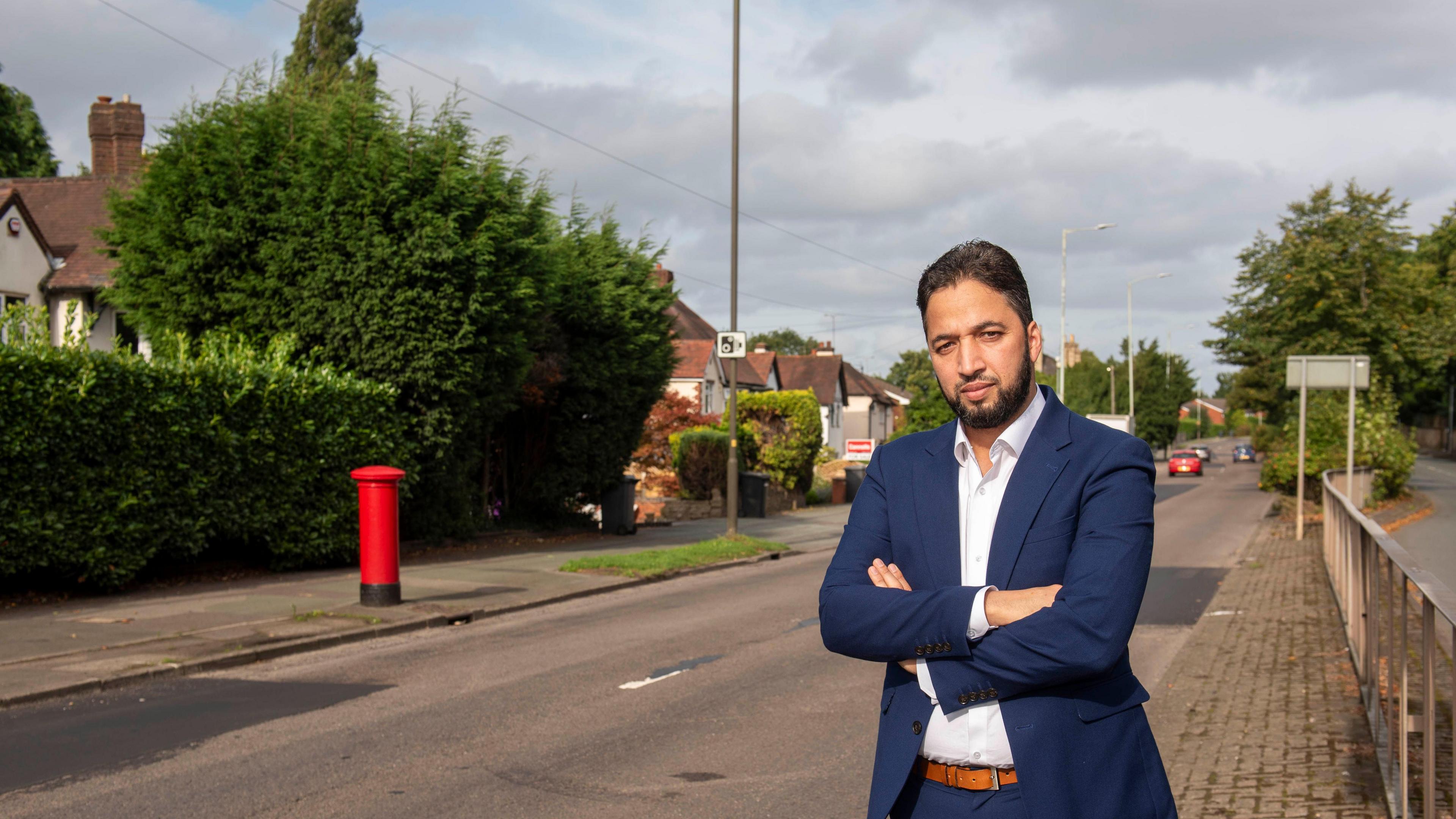 A man in a dark blue suit and a white shirt with his arms folded standing next to a road in Wolverhampton