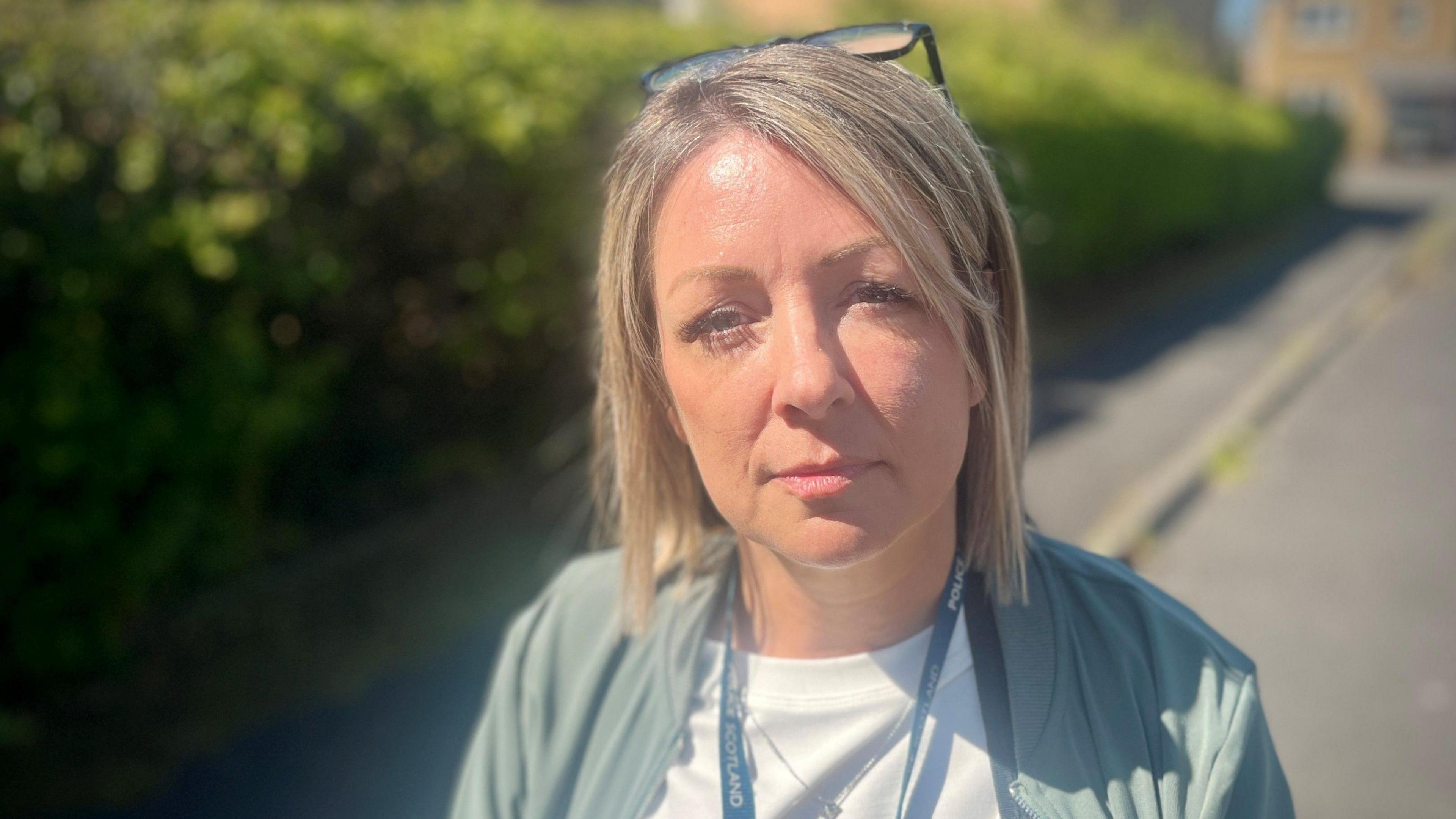 Detective Helena Scott looks into the camera, standing on the kerb with a path stretched out behind her, and a hedge. She wears a white t-shirt, a pale blue/green bomber jacket and a blonde bob with a pair of glasses on her head.