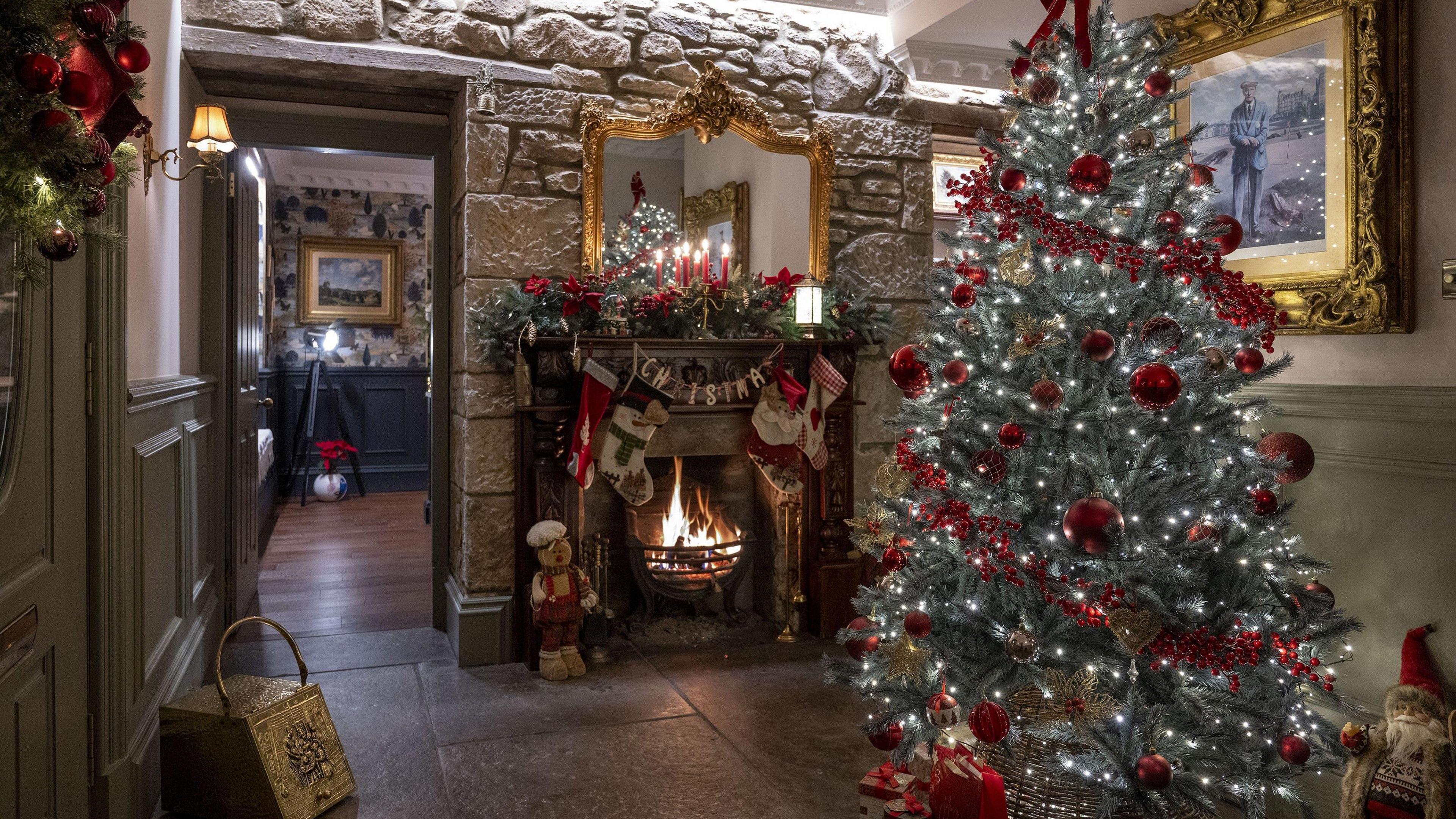 A cosy Christmas themed room with exposed brick walls. A roaring fire, adorned with stockings, Christmas foliage and candles. A gingerbread man stands to the left of the fire. A large Christmas tree, decorated with red baubles, tinsel and covered in white fairy lights sits near the fire.