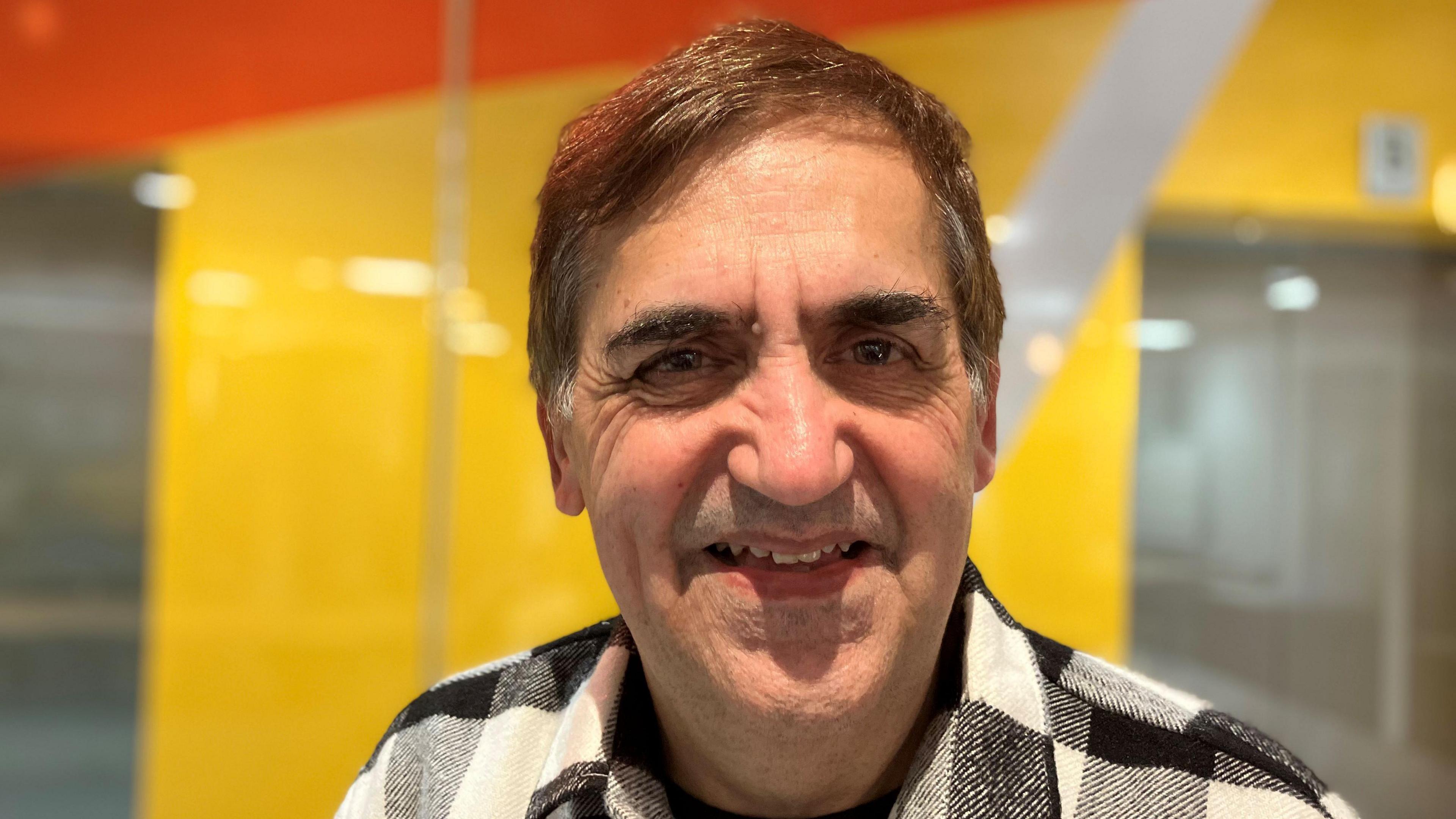 Vineet is smiling into the camera in front of a yellow background. He is wearing a black and white checked shirt over a black t-shirt. He has short brown hair.