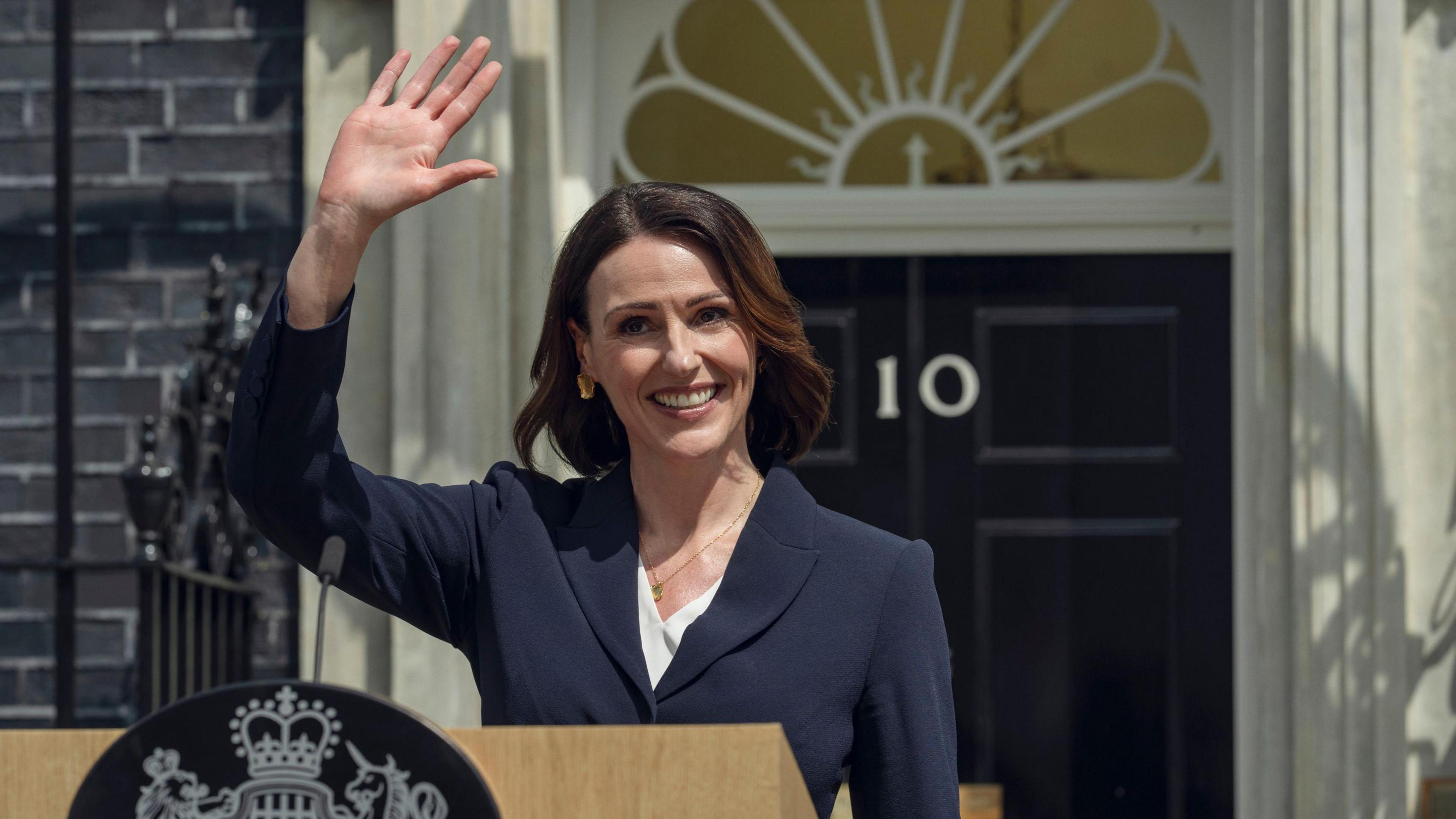 Suranne Jones playing the part of a fictional British prime minister, wearing a navy suit and waving by a podium in front of 10 Downing Street