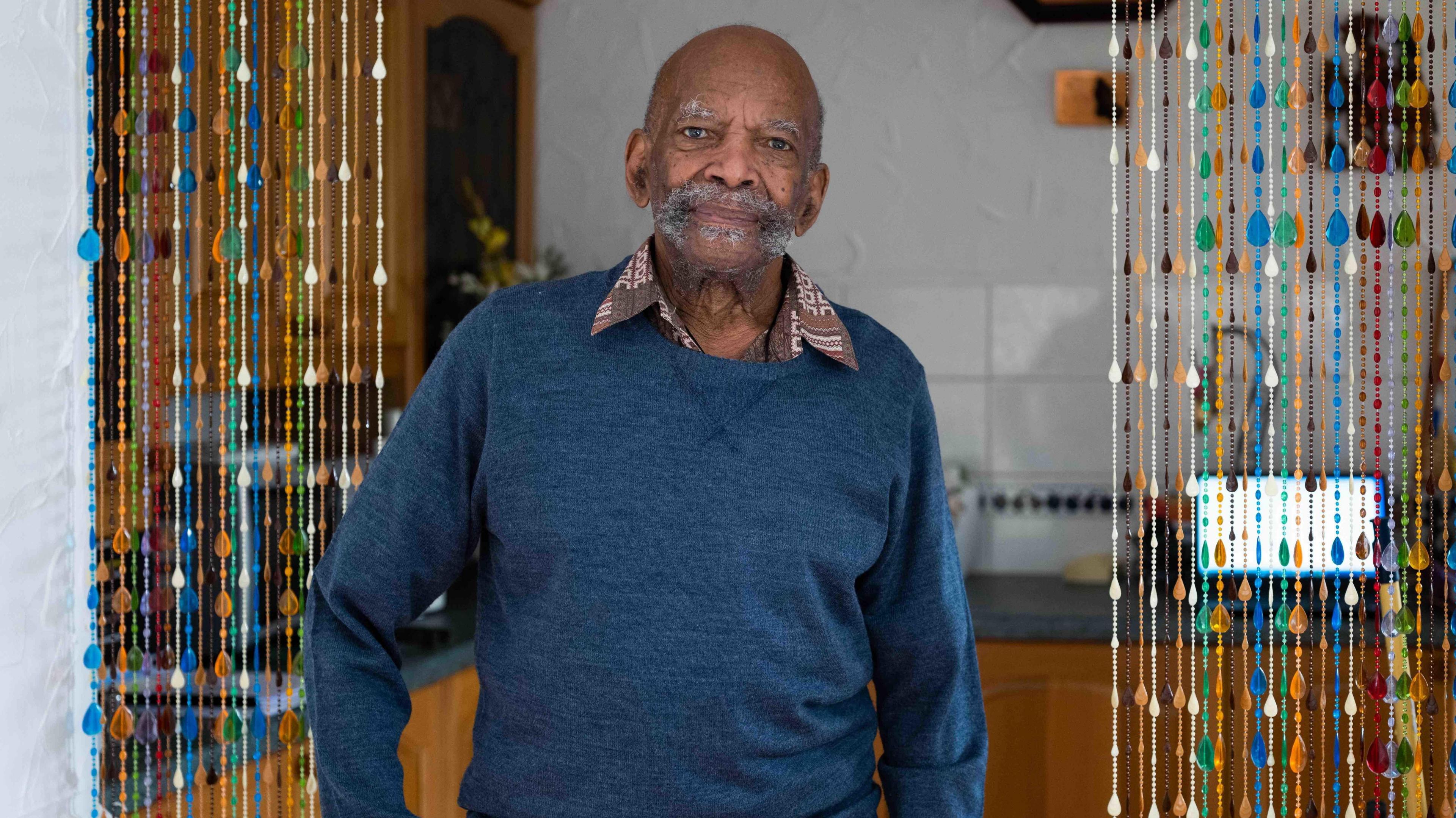 Mr Gardner, an elderly gentleman with bushy grey stubble and a blue jumper, stands in a kitchen and smiles to the camera.