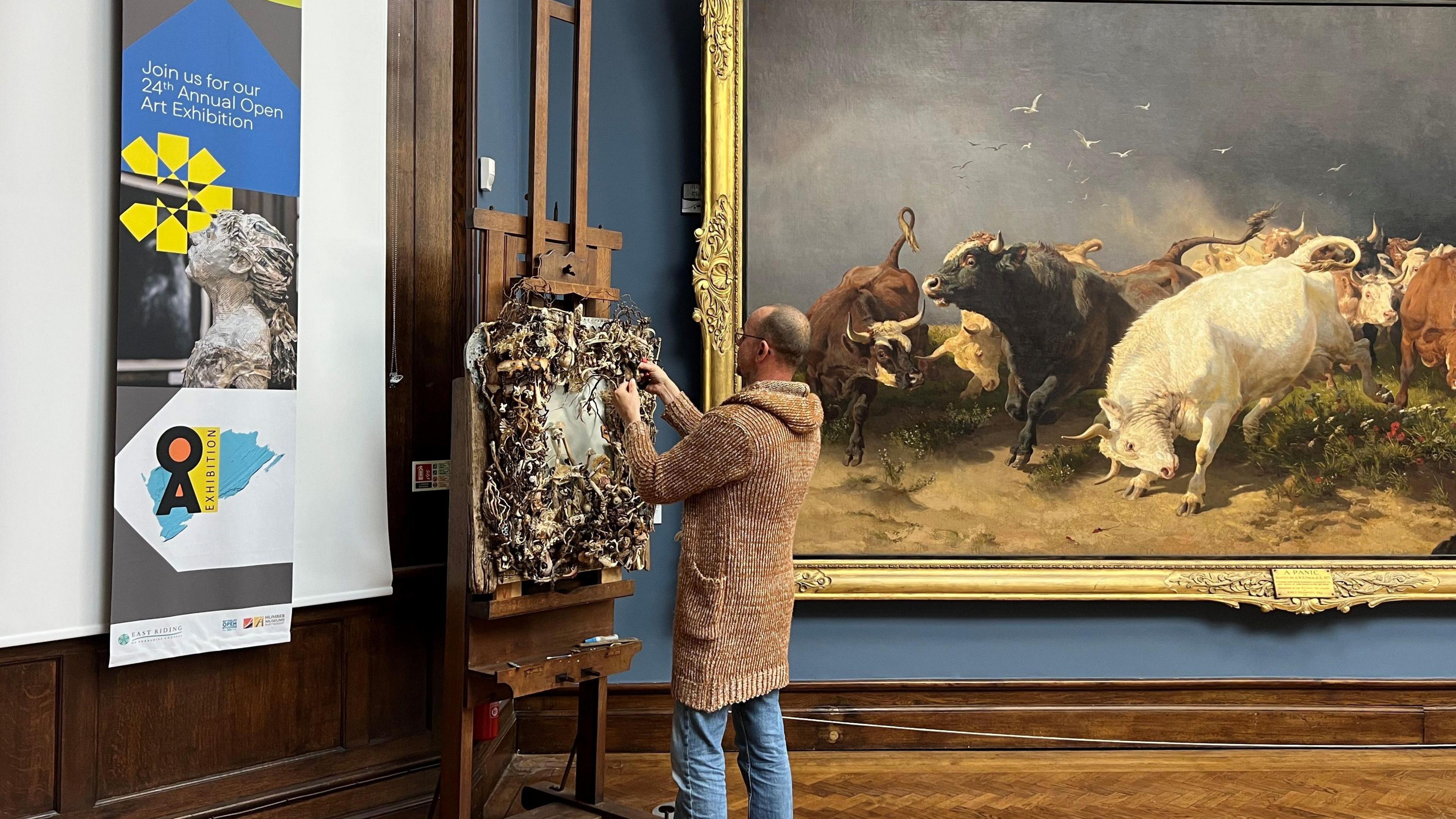 Artist Simon Baxter puts the finishing touches to his work "Life Beginnings". He is wearing a long brown jumper and jeans. His artwork is displayed next to a large ornate painting of cattle.