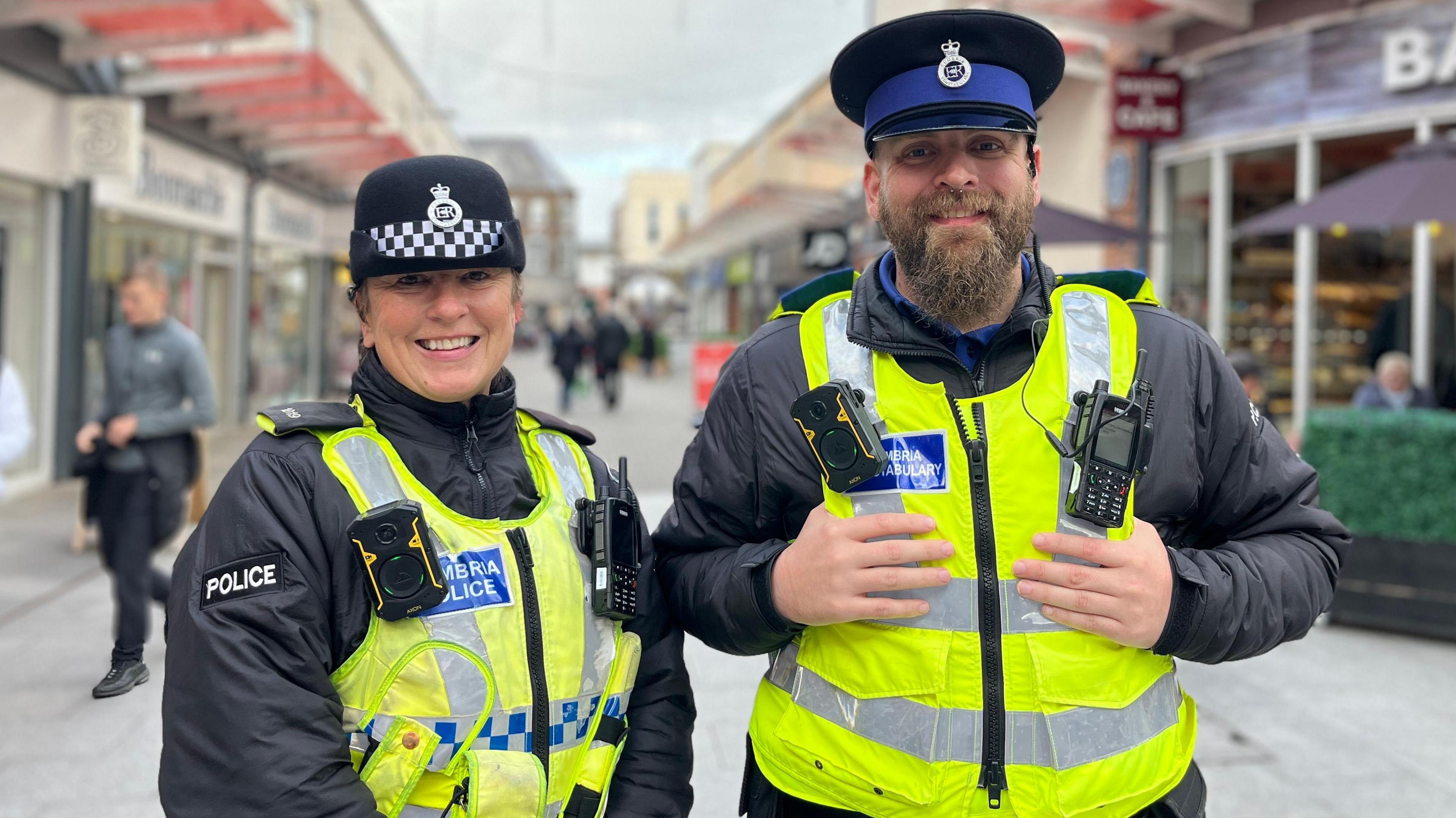 PC Jill Robertson and PCSO Dan Snell on patrol in Workington town centre. They are both in uniform, with hats and high-viz vests. They are standing next to each other and smiling at the camera. PC Robertson's hair is tied back. PCSO Snell has a beard and septum piercing.