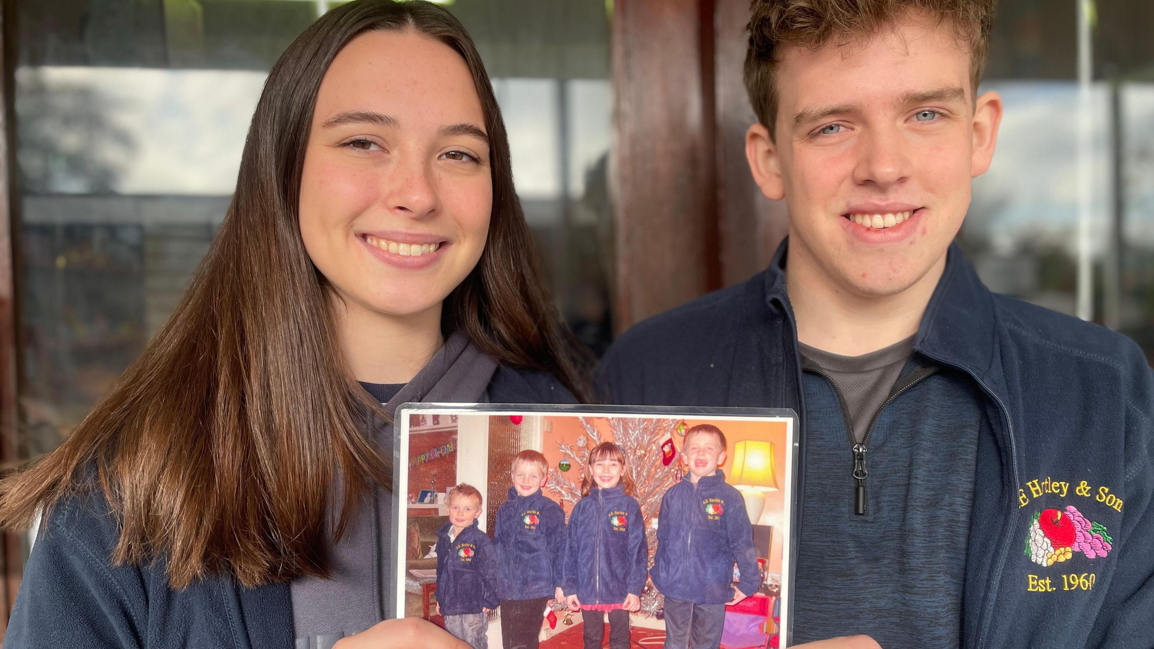 A young woman with long brown hair smiles while holding up a picture. The boy next to her also smiles. Both wear blue fleeces.