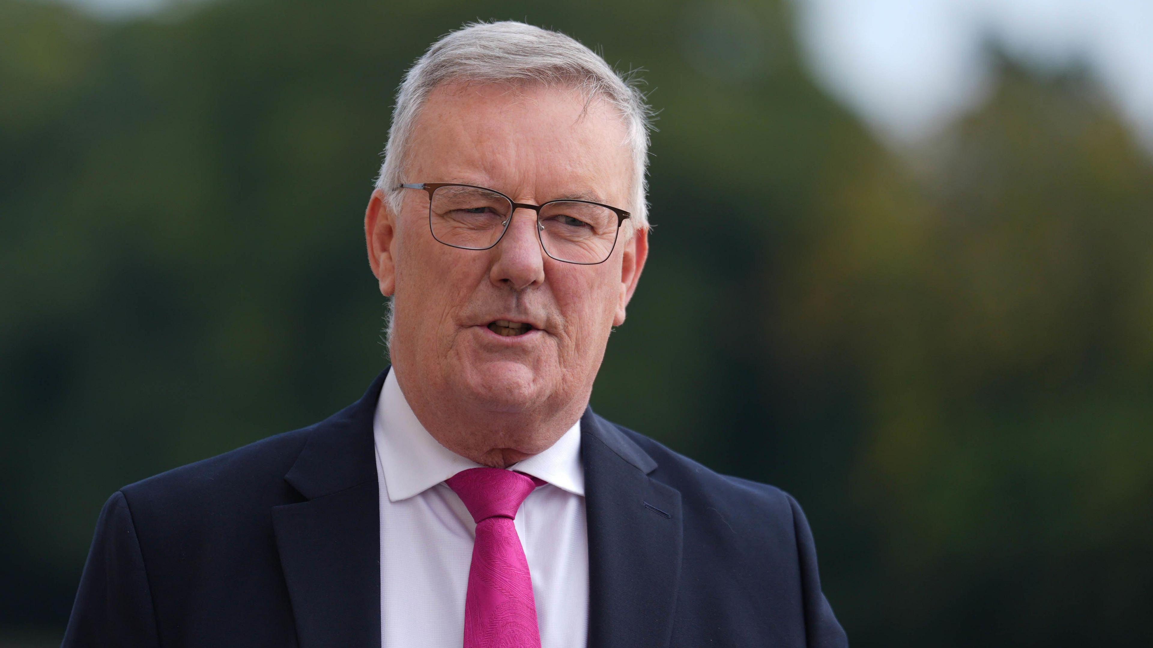 Mike Nesbitt wearing a dark suit, white shirt, and pink tie. He is wearing dark rimmed glasses and has grey hair. 