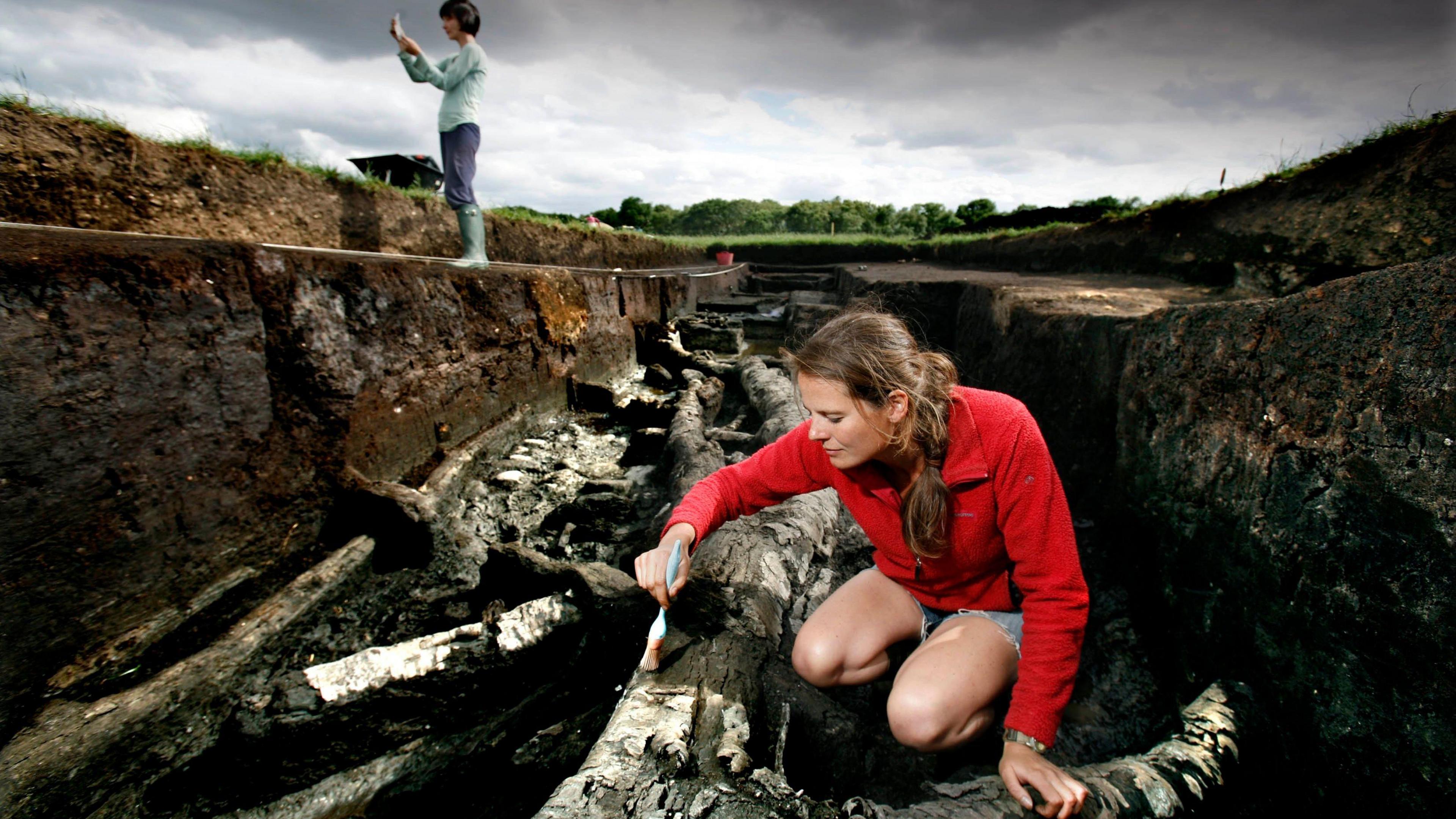 A woman in a red jacket brushes a stone wall in an archaeological site