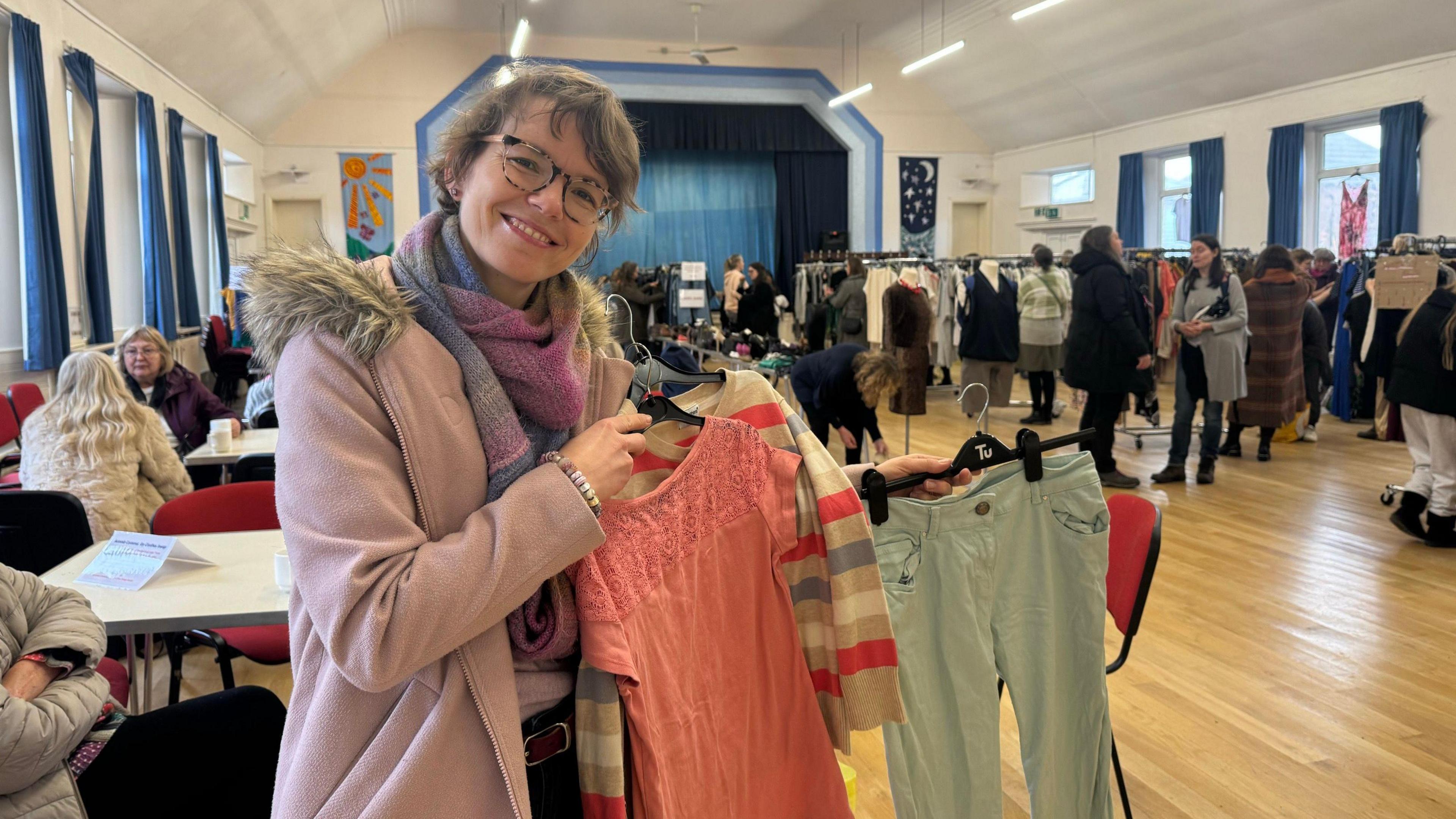 A woman in a purple coat holds up three hangers with trousers, a cardigan and top on. She stands in a church hall set up with clothes rails.