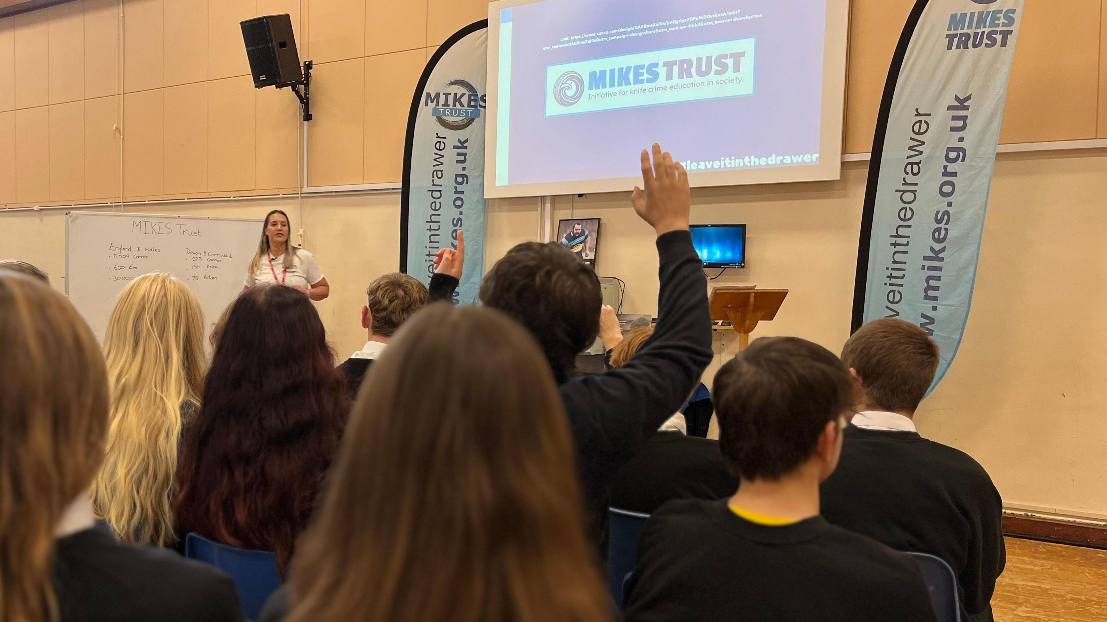 A woman in a white t-shirt stands in front of a class of year 10 students in a school hall. Behind her is a presentation screen with a logo for MIKES TRUST. In the foreground one of the students is raising their hand to ask a question. 