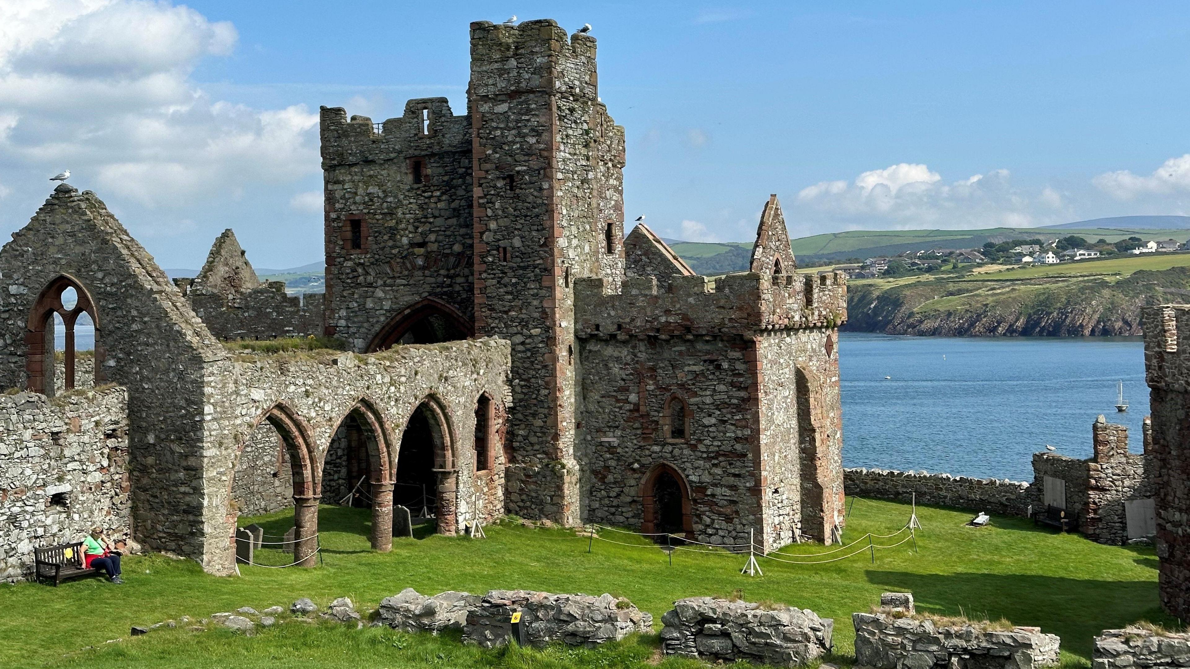 A large part of the ruins of Peel Castle, which is built in sandstone and is surrounded by green grass with Peel Bay in the background with a headland above it.