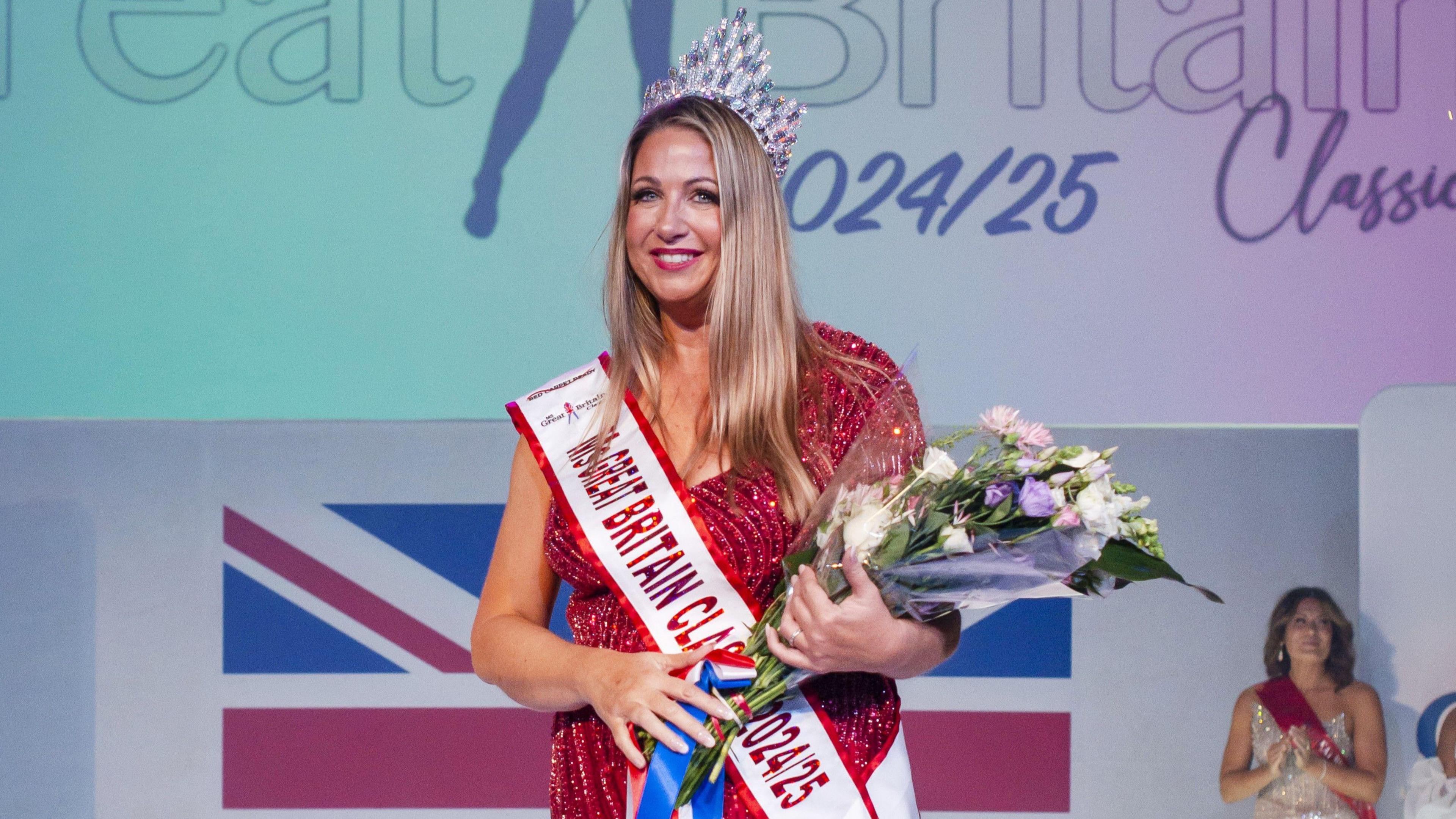 A front view of Debbie Hughes. She is wearing a sparkly red long dress and a silver crown on her head. She has a sash on that says "Ms Great Britain Classic", and she is holding a bouquet of flowers, and has the other hand on the crown. She has long blonde hair