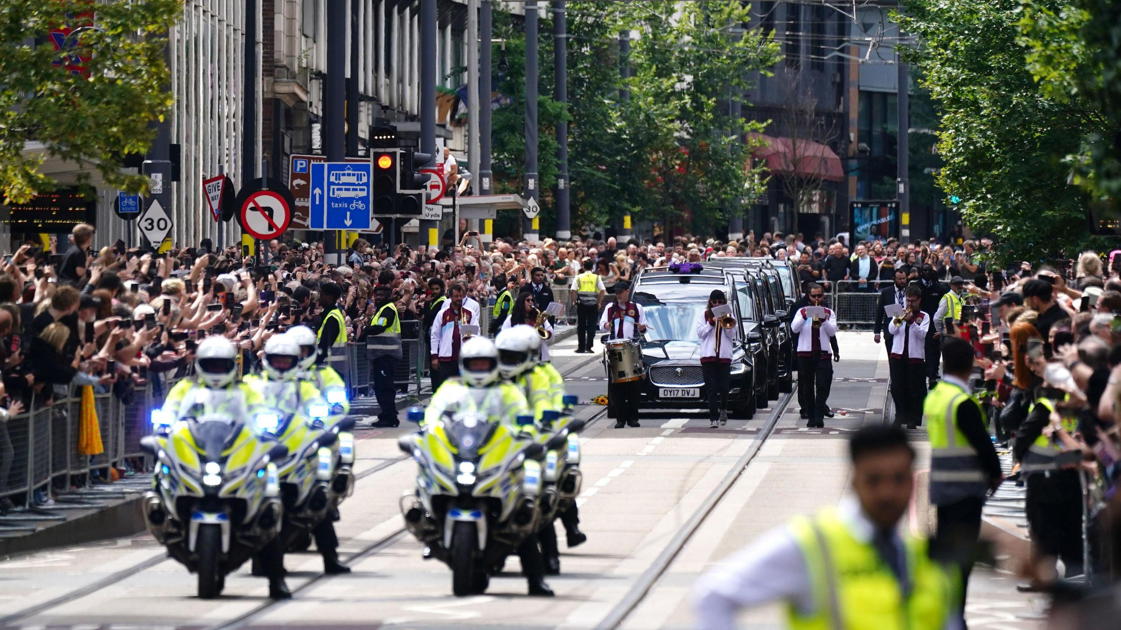 Thousands of people line either side of a street behind metal barriers, holding up their phones. A police escort of six motorbikes drive in front of a line of black funeral cars. The front car has purple flowers on its roof.