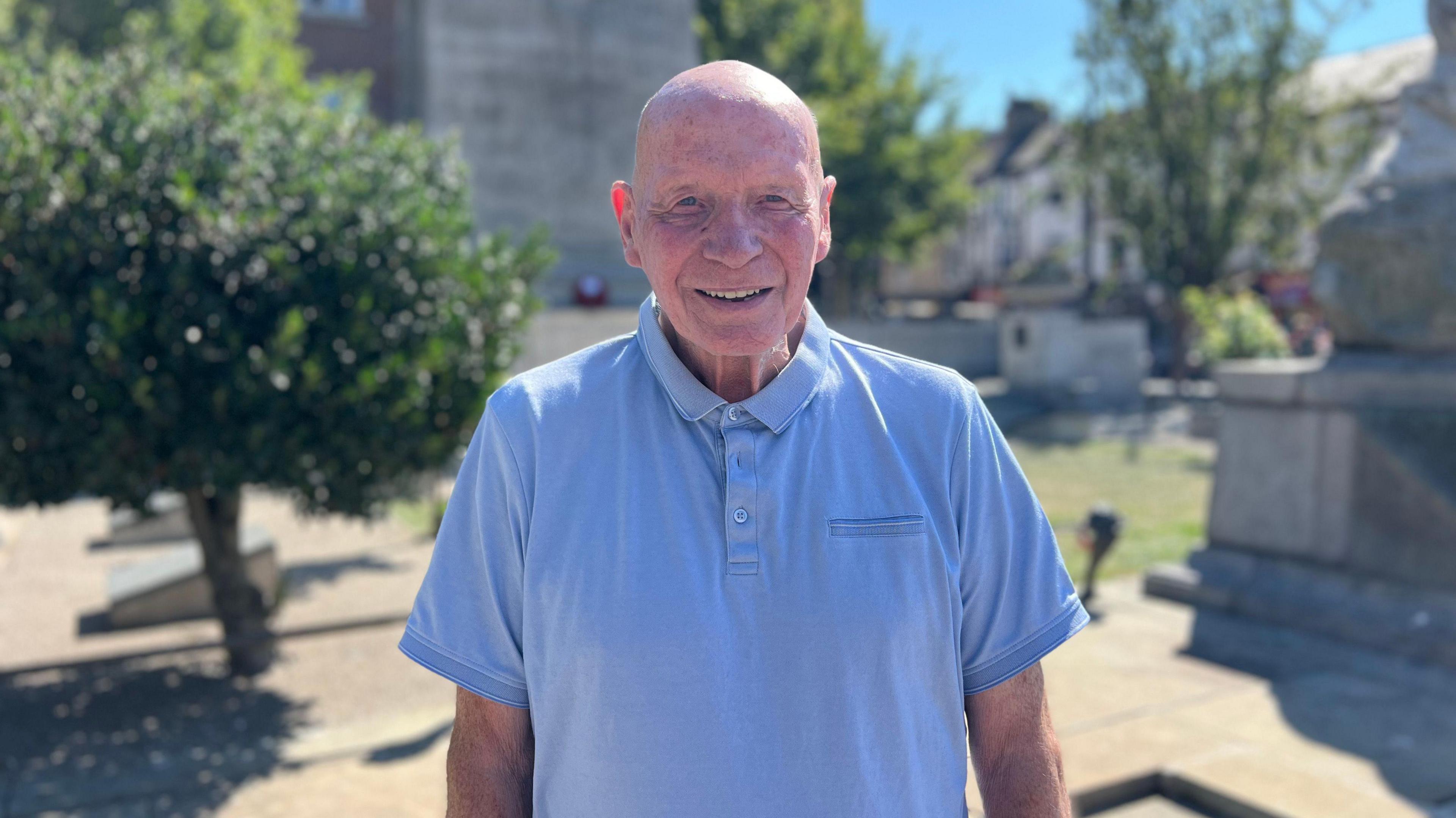 A man with a bald head and a light blue polo shirt, smiling at the camera. He is standing outdoors on a sunny day with trees and a monument in the background.