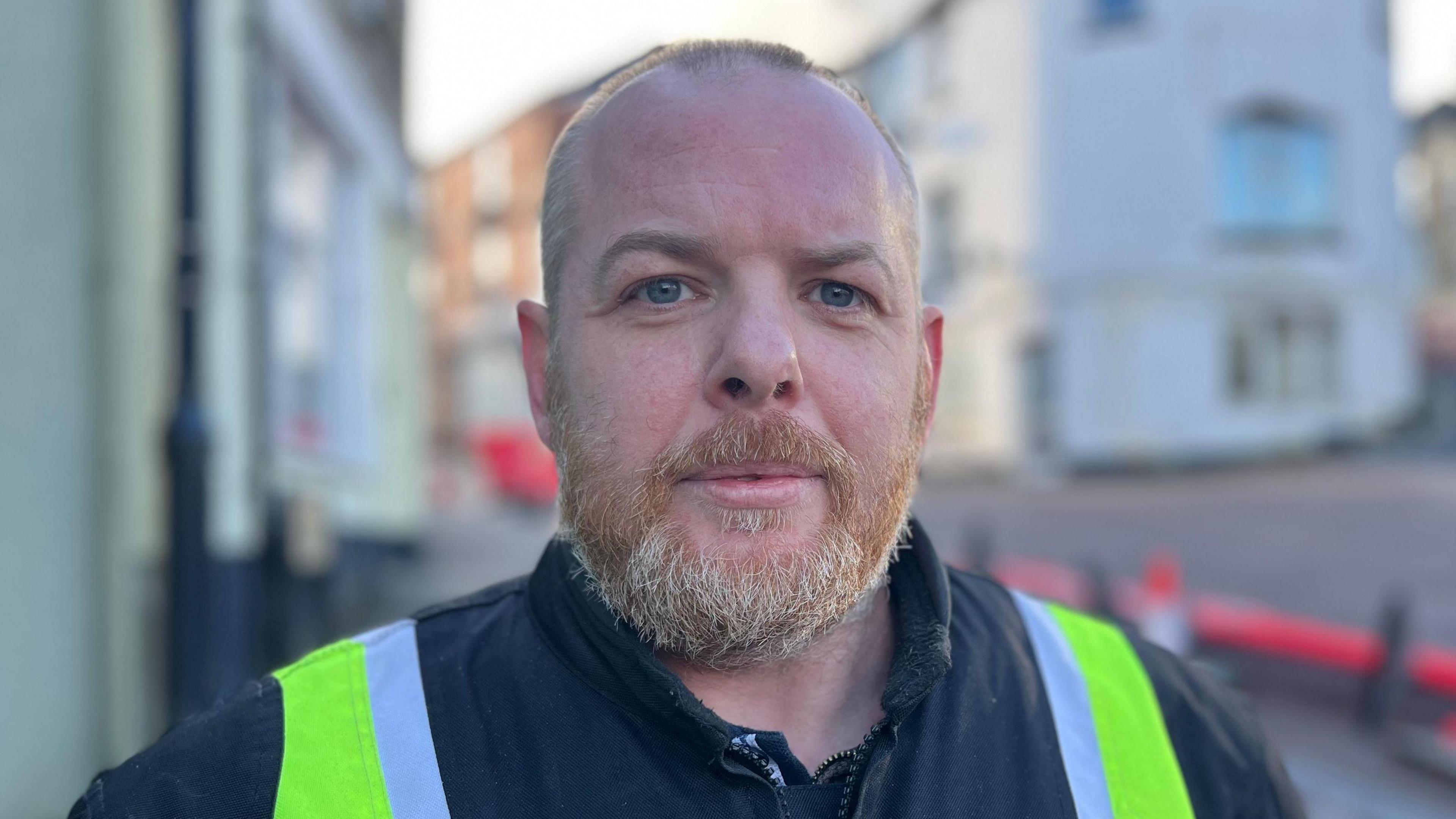 Adam Brophy stands next to a section of road in Bodmin which has been closed off while an unsafe building which is over his left shoulder is examined. He is wearing a black jacket with a high-vis jacket over the top of it. He has short hair and a ginger and grey beard.