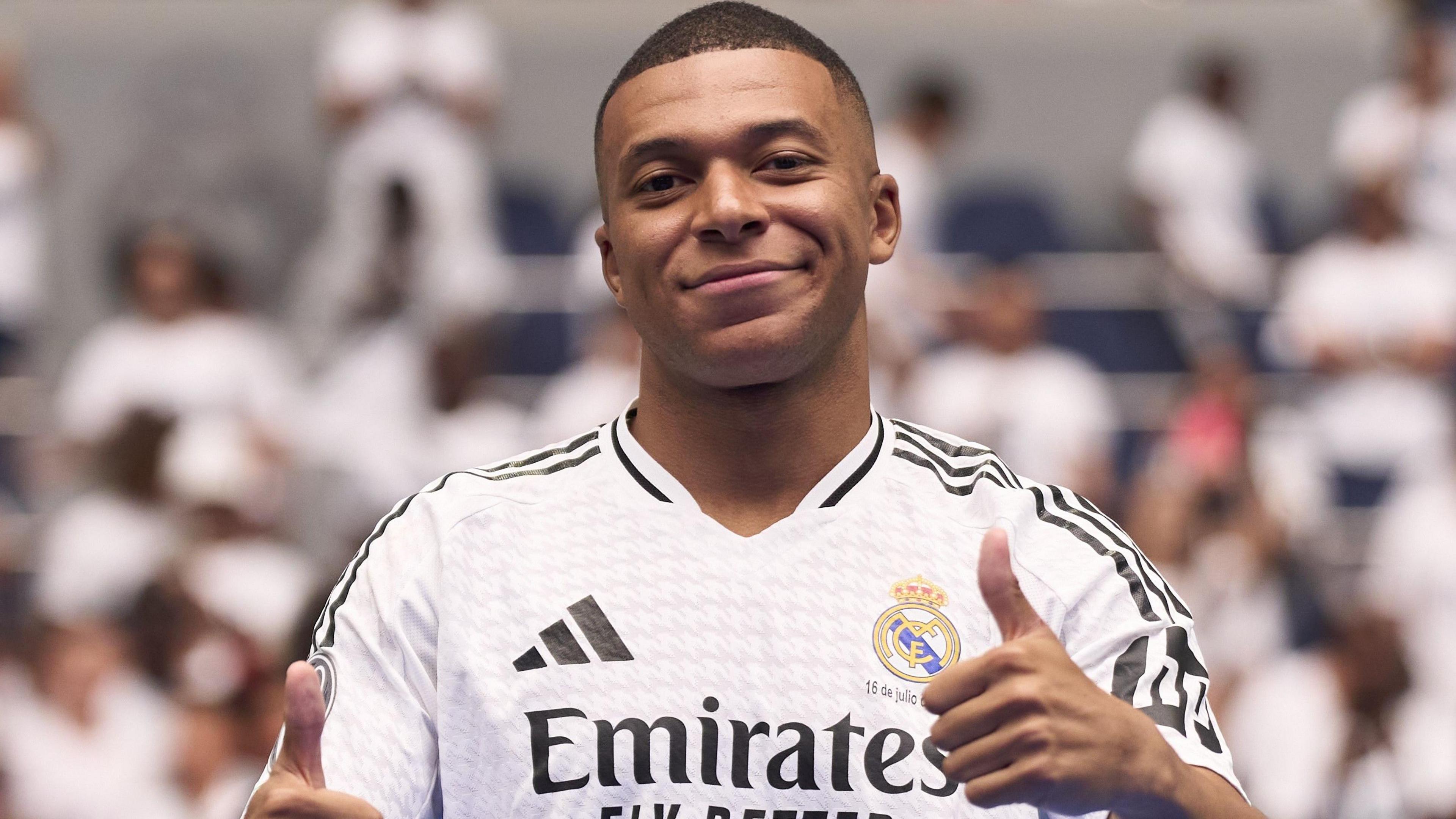 Kylian Mbappe wearing Real Madrid's white jersey with his thumbs up. There is a blurred background of fans