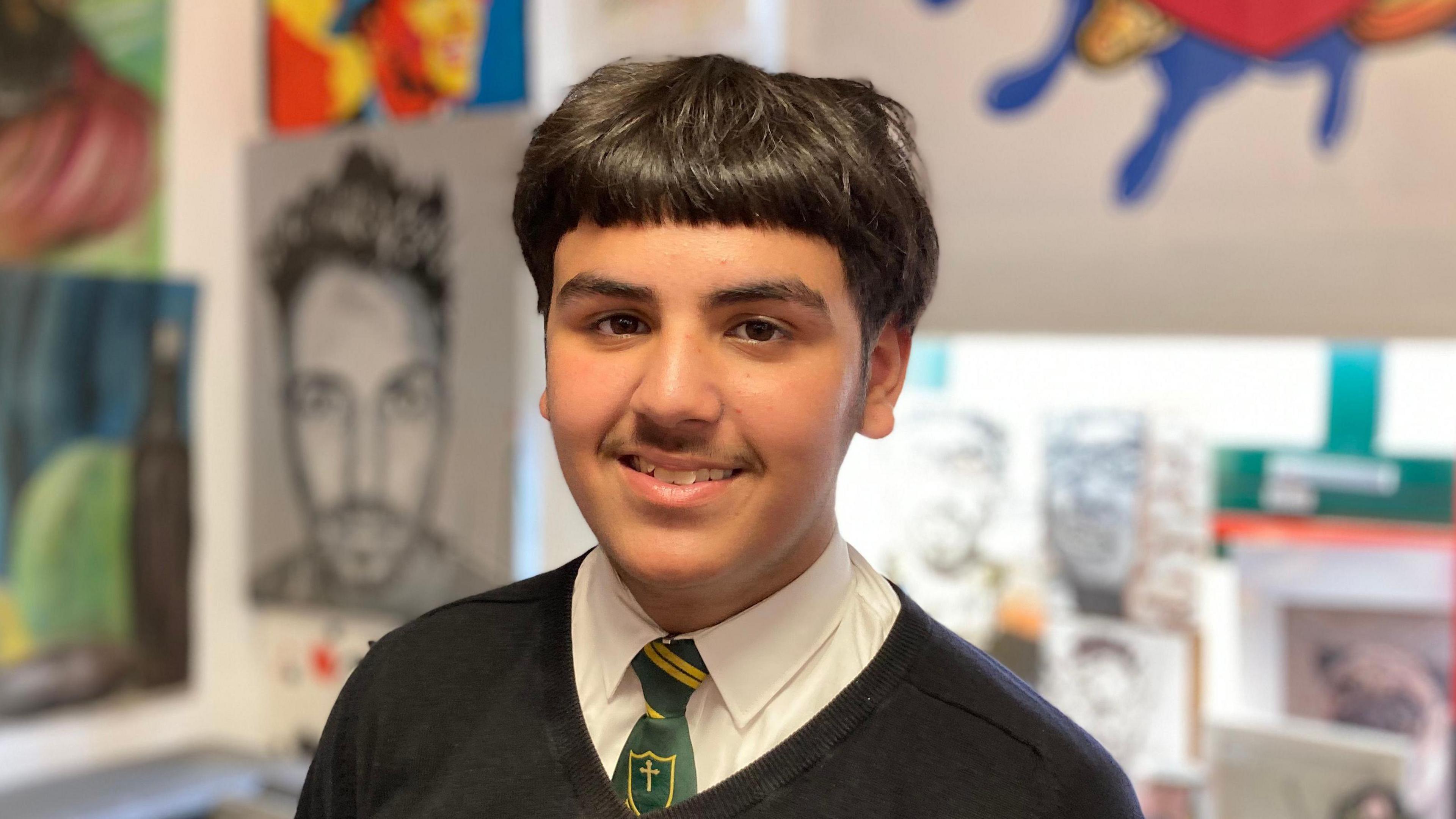 A teenager of South Asian heritage smiles toward the camera, he is wearing a dark green tie, white shirt and black jumper, behind him are pieces of hand drawn artwork on the wall.