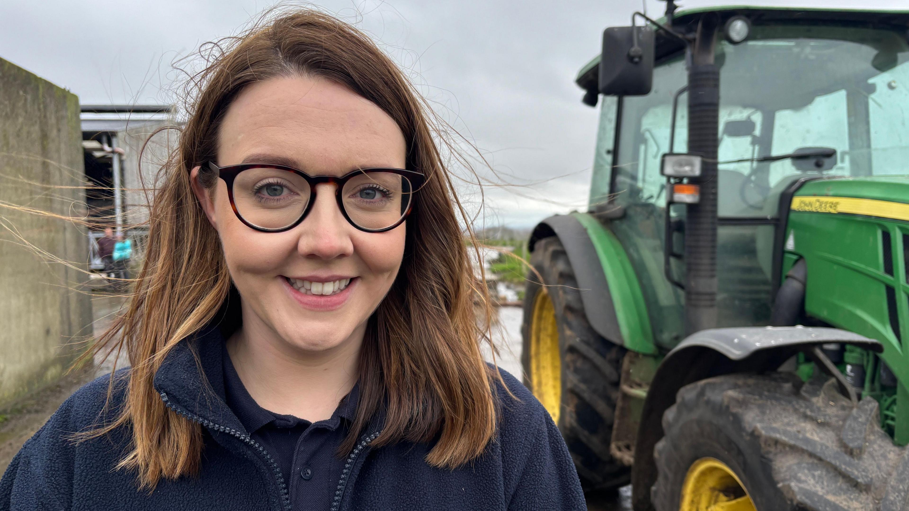 A young woman stands in a farmyard. She has auburn shoulder length hair which is being blown by the wind. She's wearing glasses and smiling. Behind her is a green tractor and grey skies.