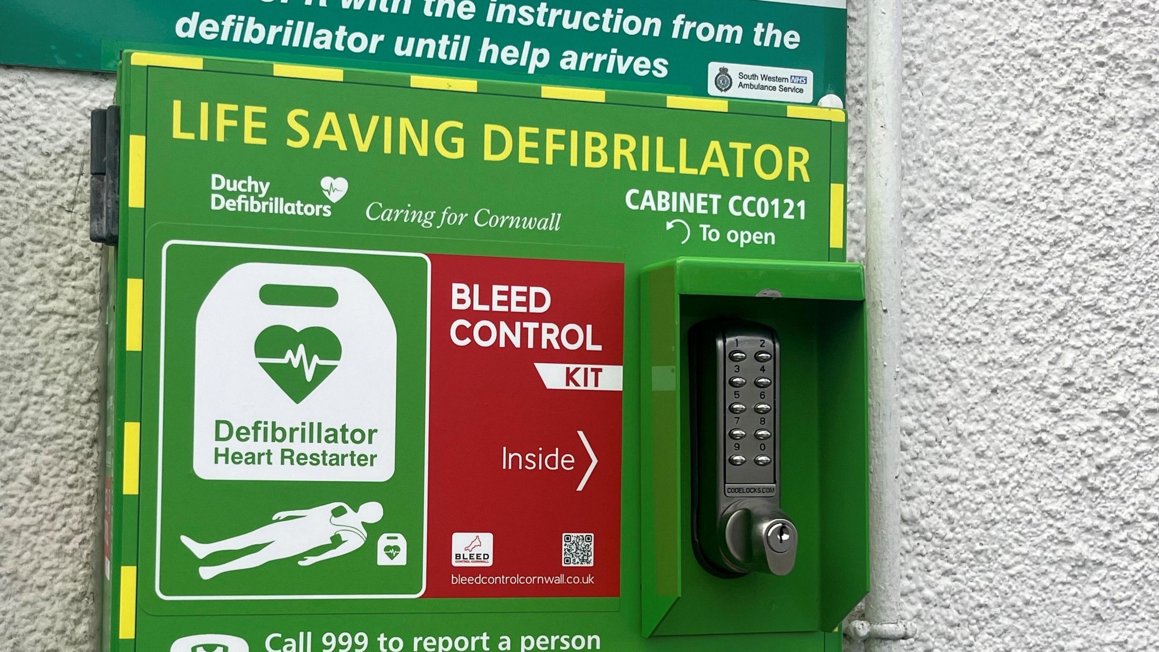 A green cabinet on a white wall with a security handle to open, it is covered in lettering which explains it is a life saving defibrillator from Duchy Defibrillators. It also has a red sticker on it which says bleed control kit inside.