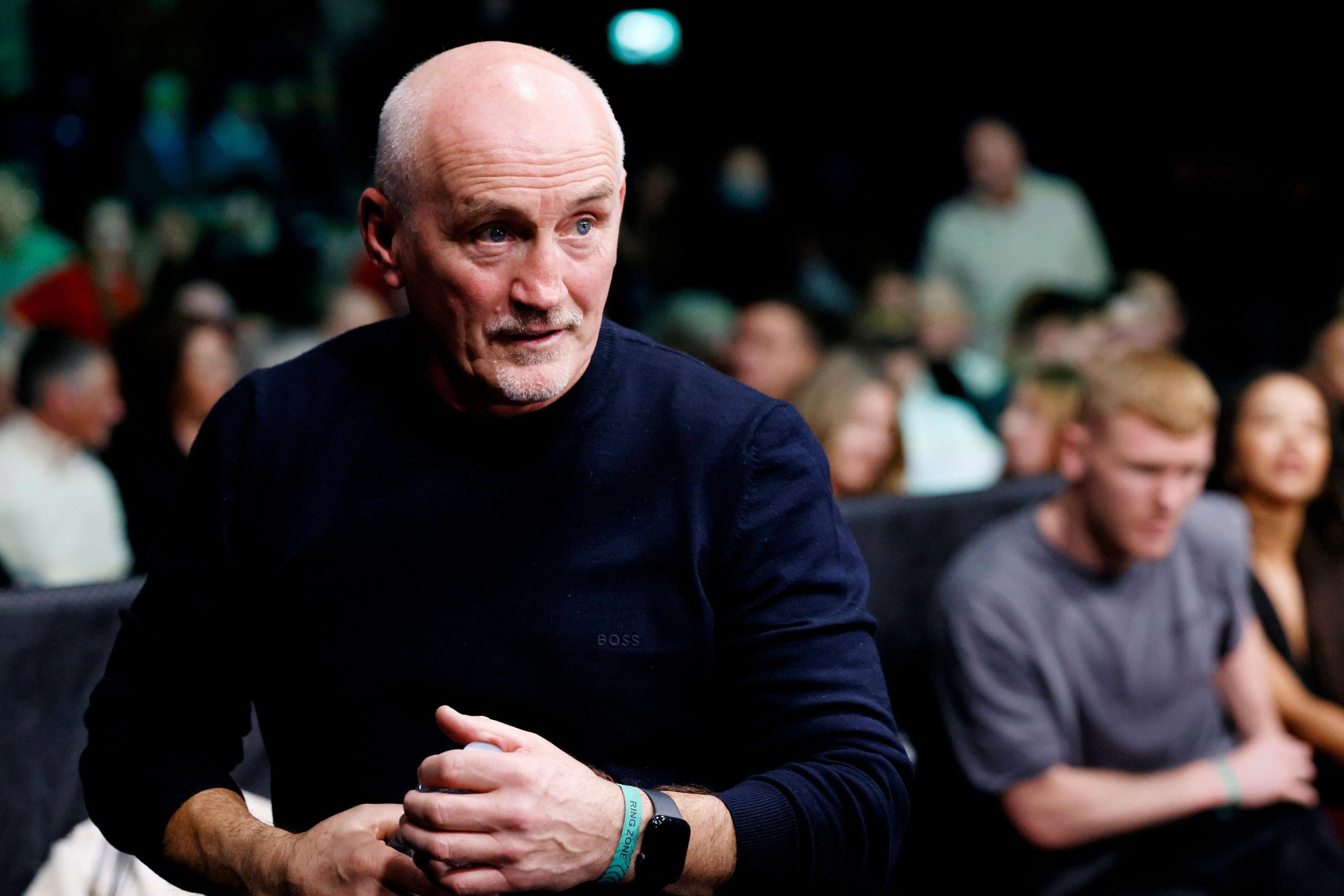 Barry McGuigan watches a boxing match at ringside. He has a bald head, a grey beard and is wearing a blue jumper with the sleeves rolled up. A large crowd is visible behind him.