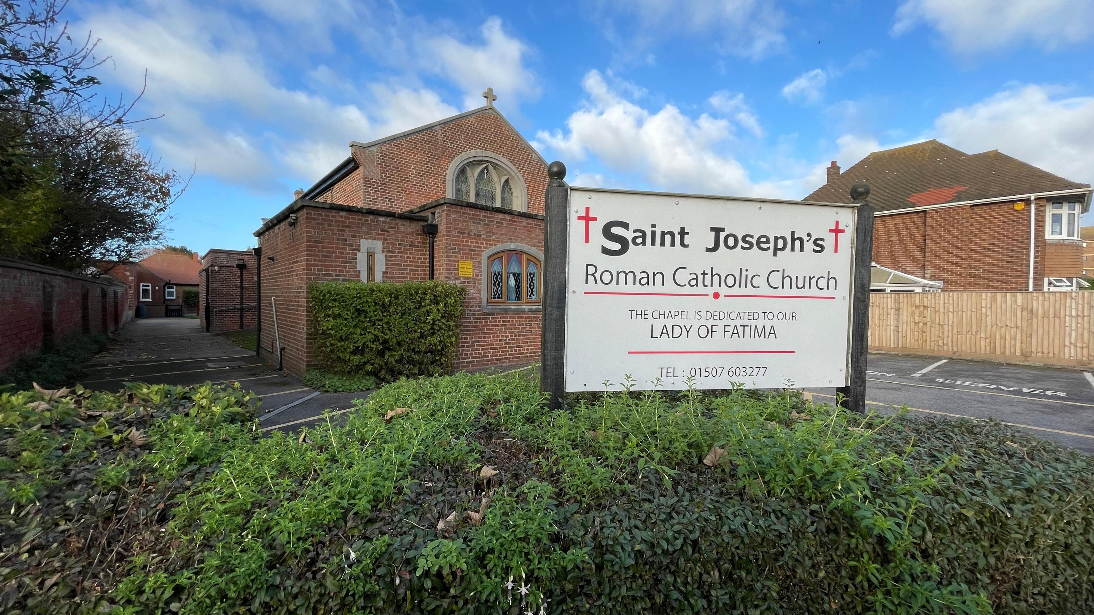 A view of a church with a bush and car park in the foreground and a sign saying Saint Joseph's Roman Catholic Church with crosses either side.