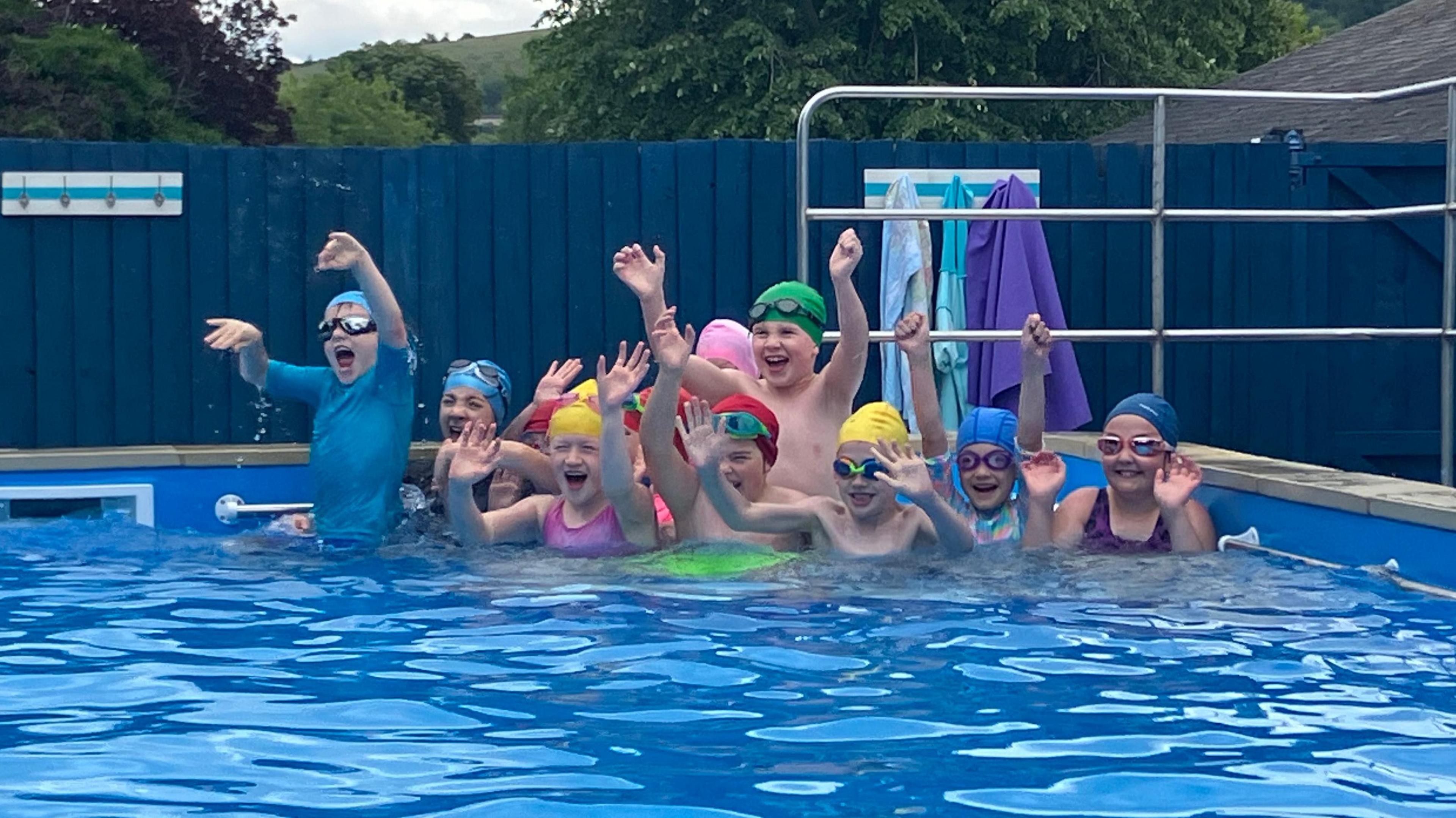 A group of school children, all wearing multicoloured swimming costumes, hats and goggles cheer with their arms up in a swimming pool.