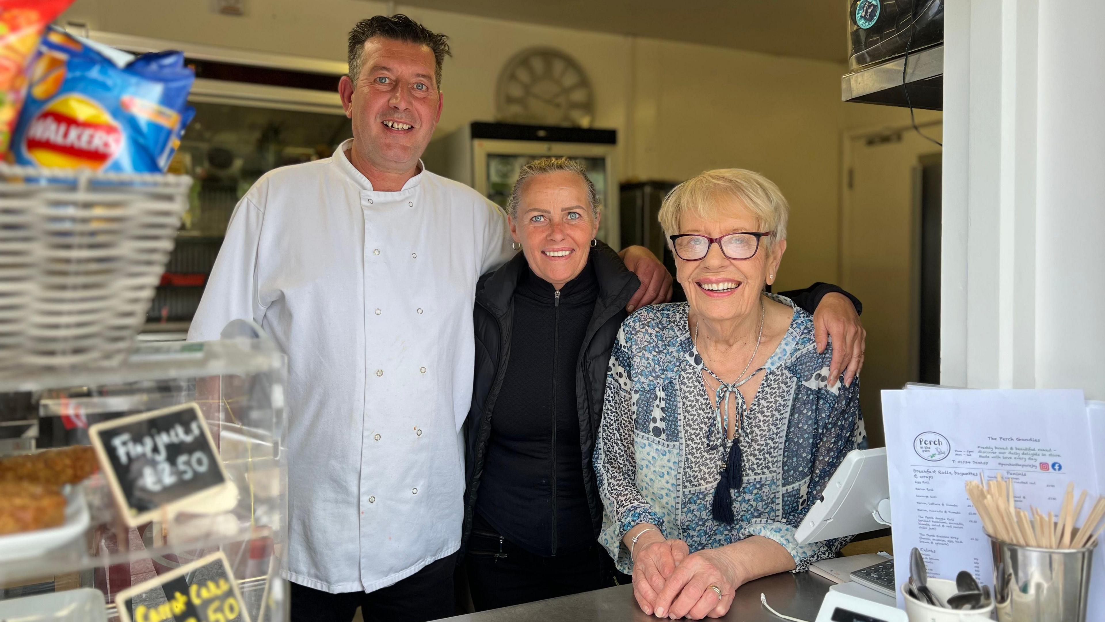 Dawn Kennedy is in standing in a kiosk with her arms around an older woman on the right and a taller man in a chef jacket to the left.