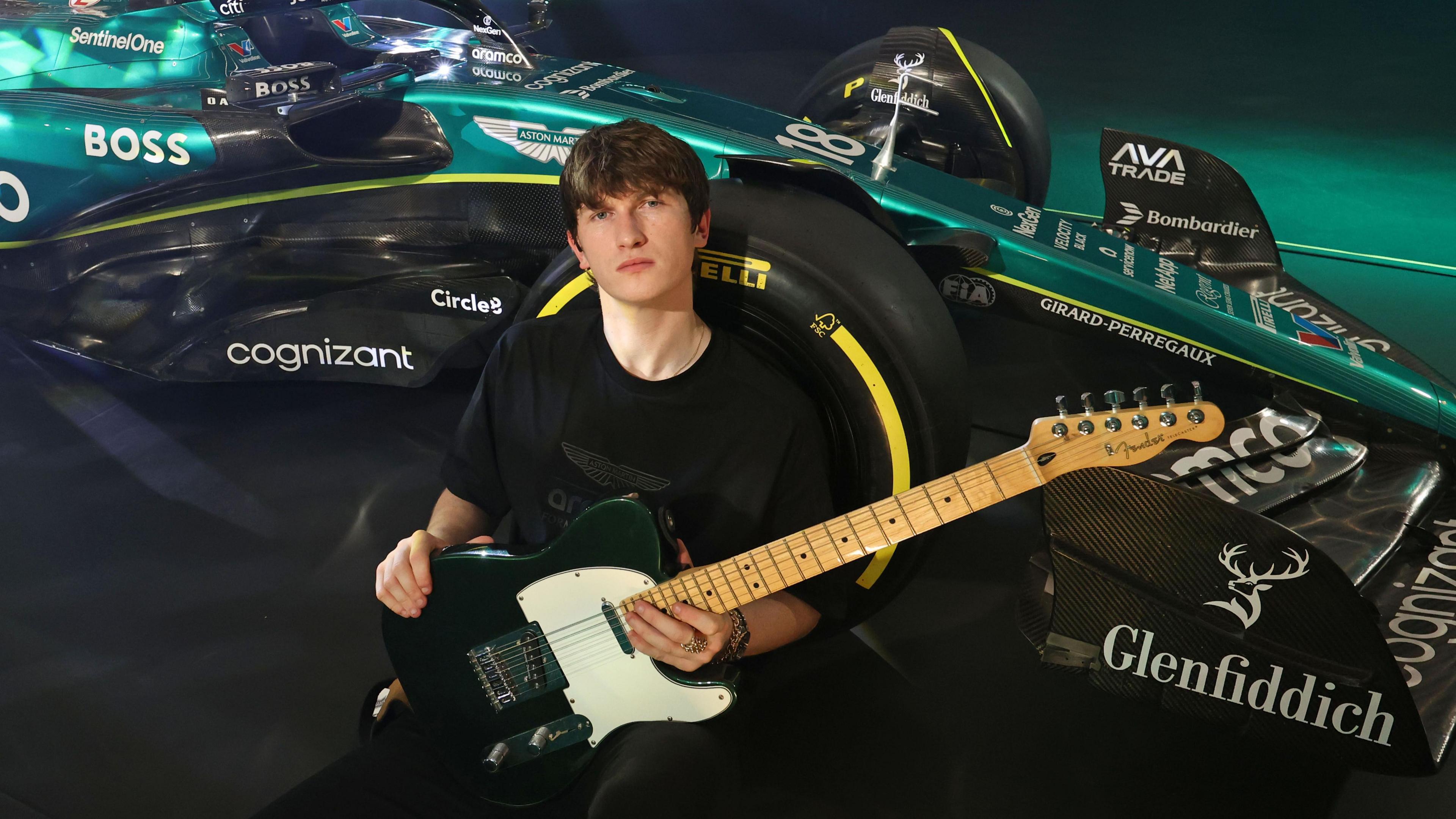 A young man wearing a black top and holding a black and white guitar leaning up against a green British formula one car 