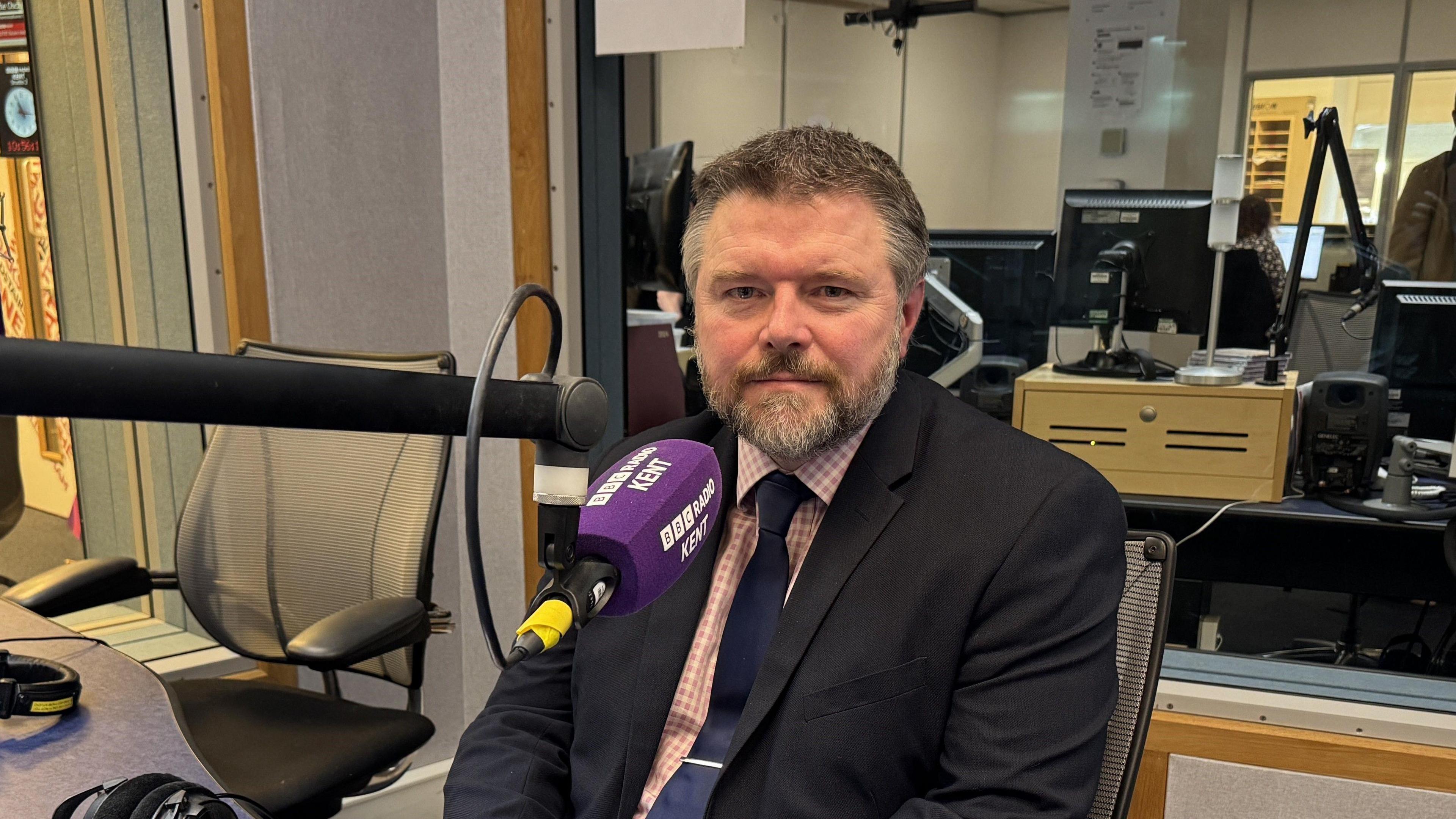 A man with a beard and wearing a suit sits in a studio with a BBC Radio Kent microphone on front of him.