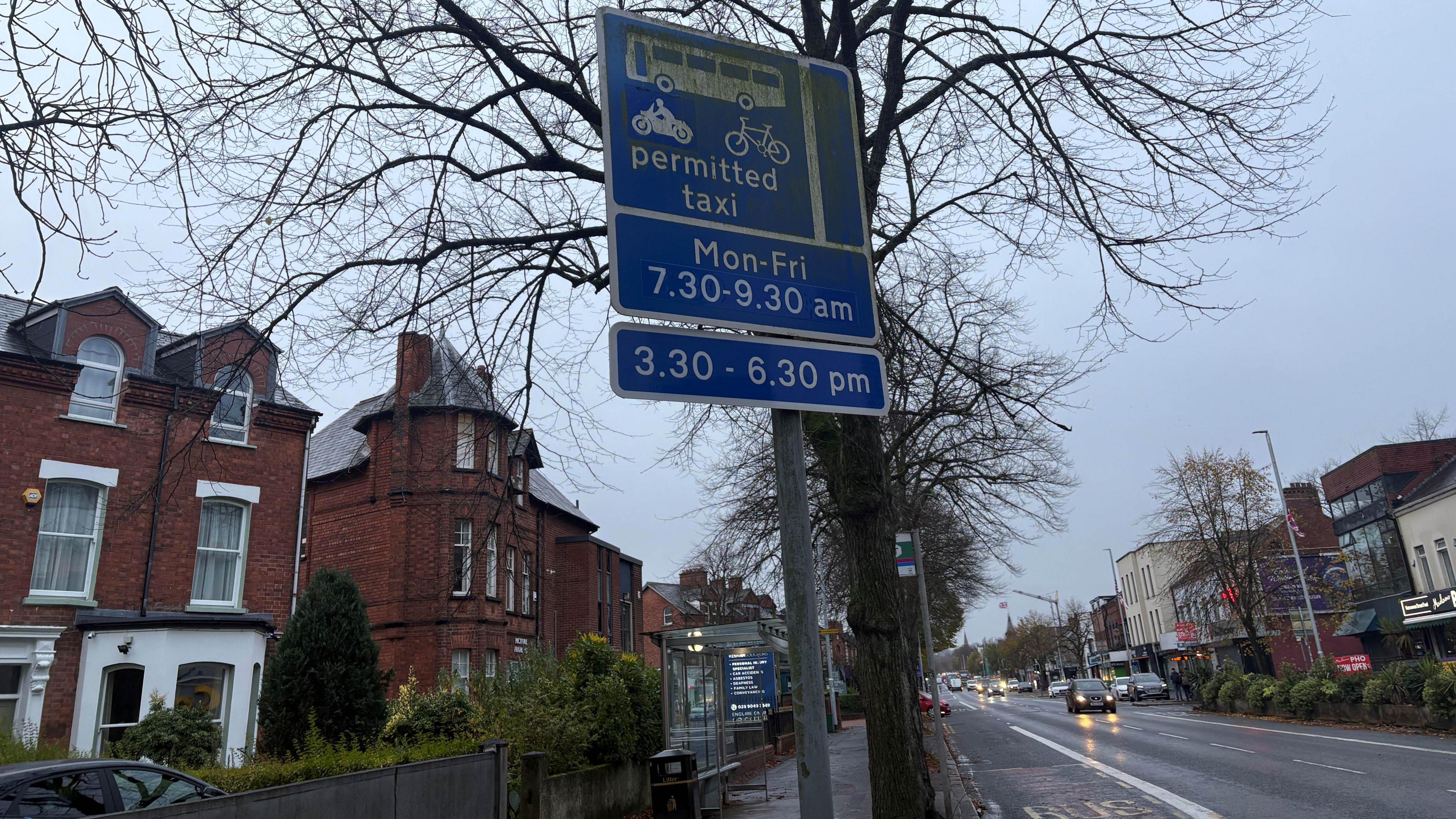 A blue sign with white writing reminding drivers of the new bus lane times. It states: permitted taxi. Mon-Fri, 7.30-9.30am. 3.30-6.30pm. Houses are in the background. 