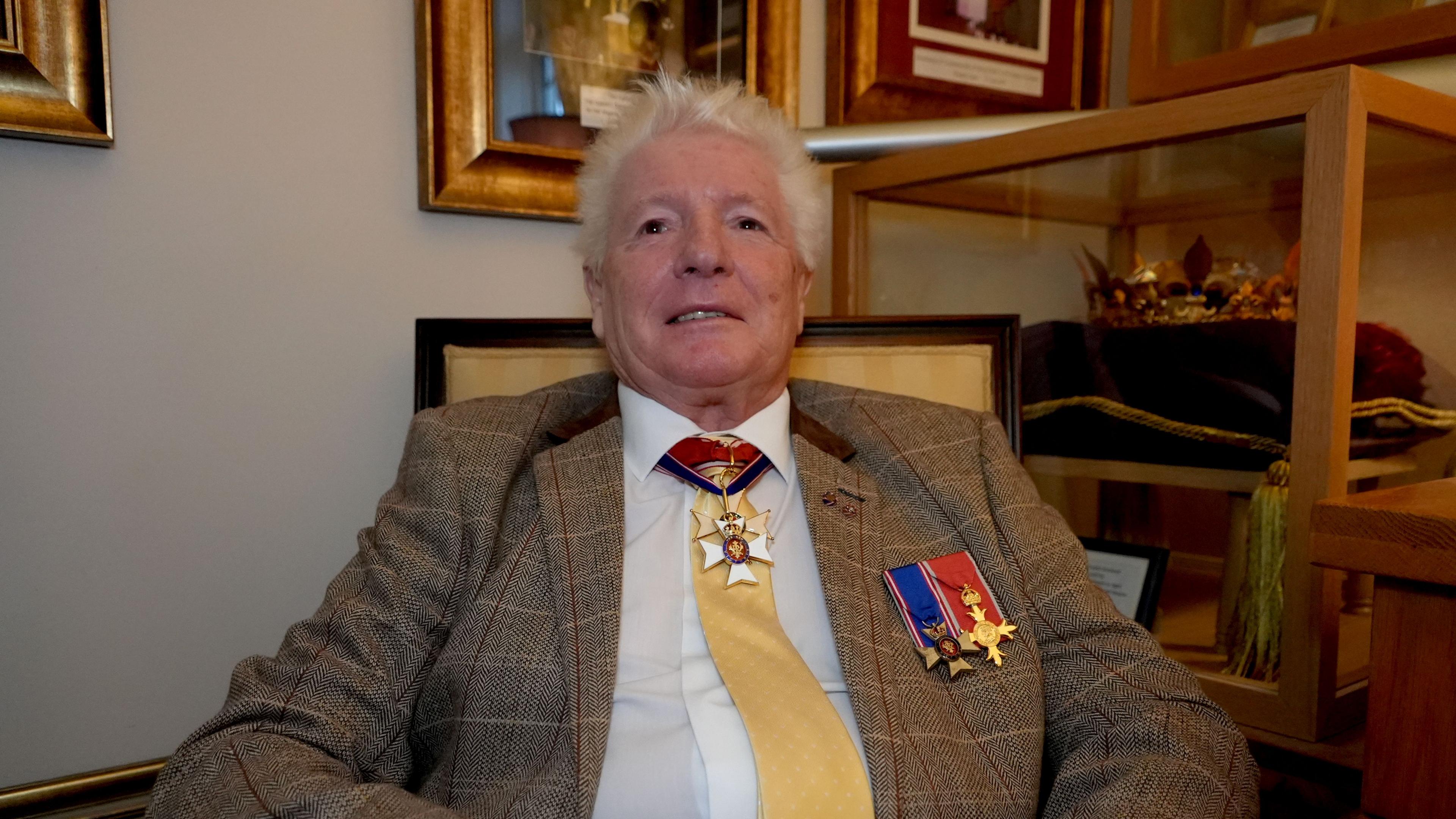 Bruno Peek sits in an armchair. He is wearing a white shirt and tweed jacket. Two medals adorn his jacket with one around his neck rests on his gold tie. There are gold-framed photos behind him on a wall, and a glass cabinet contains a crown.