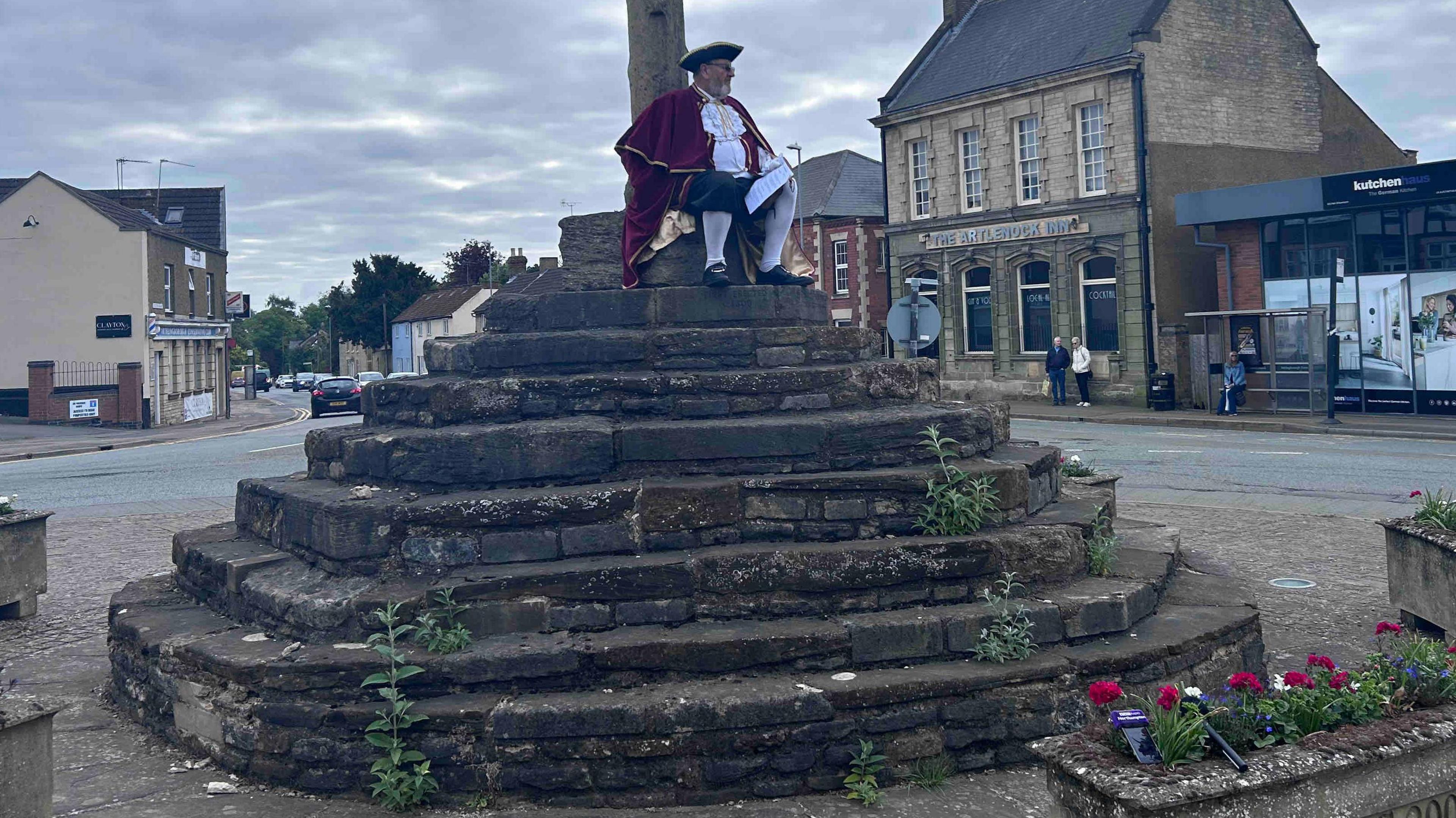 Stone steps in the centre of a town - there are weeds growing from some steps. A town crier in red and white with a black tricorn hat is sitting on the top step. There are shops and houses in the background, and a stone planter in the foreground.