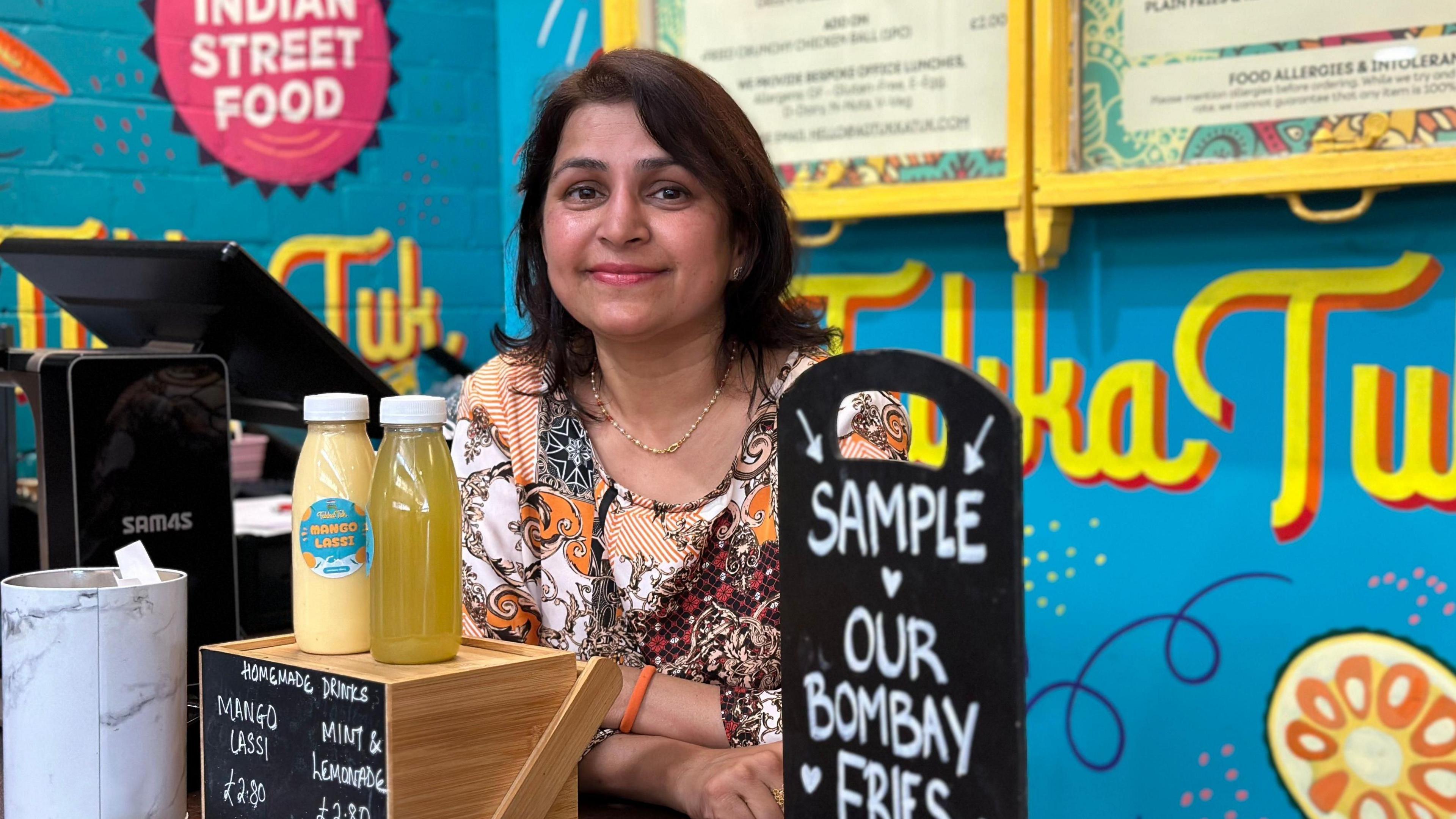A woman with brown hair stood behind a food counter, the signs in the background are bright blue, with the word Tukka Tuk written in bright yellow. The woman is wearing an orange and white top.