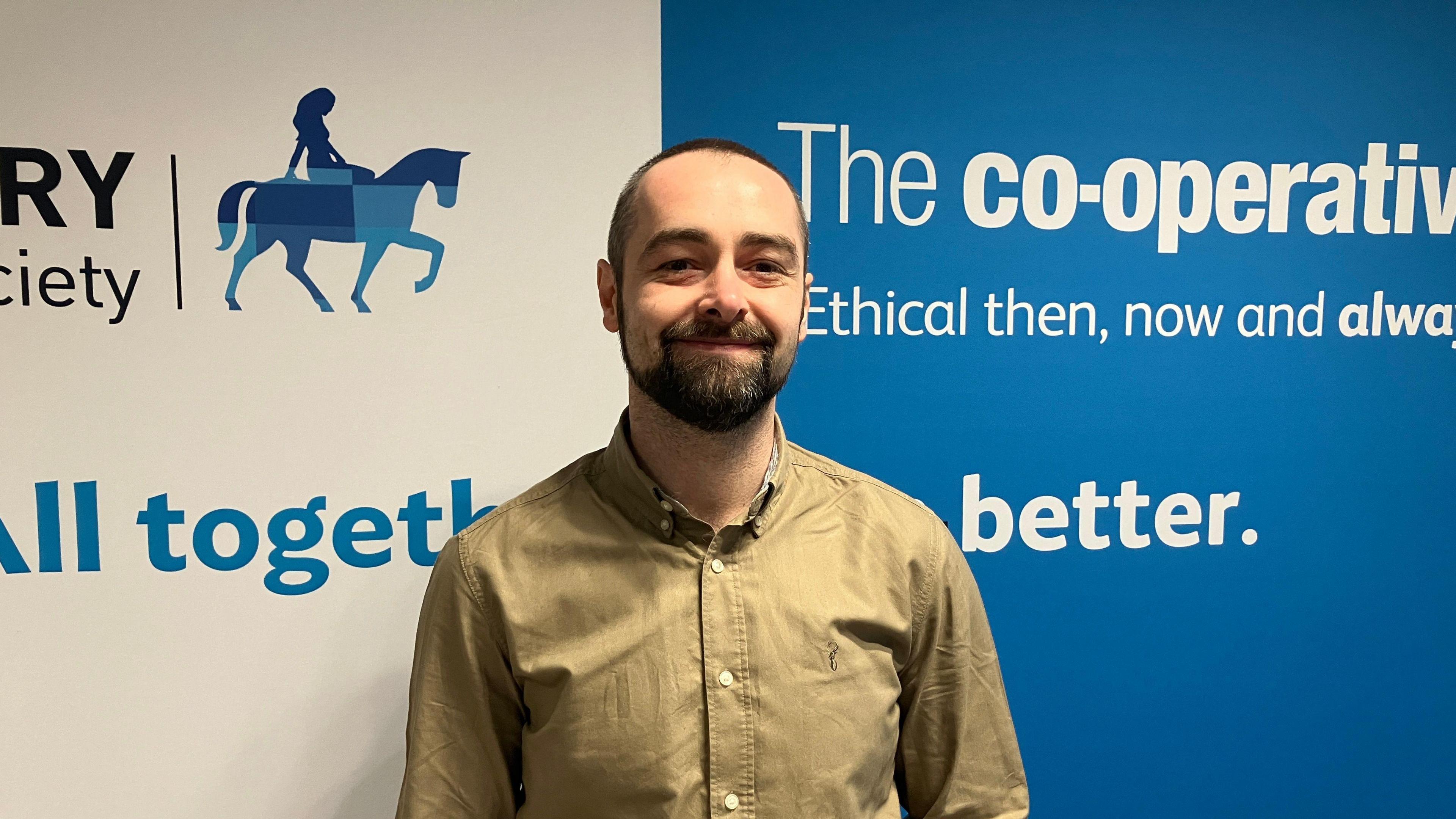 A man in a beige shirt smiles in front of a blue and white banner.