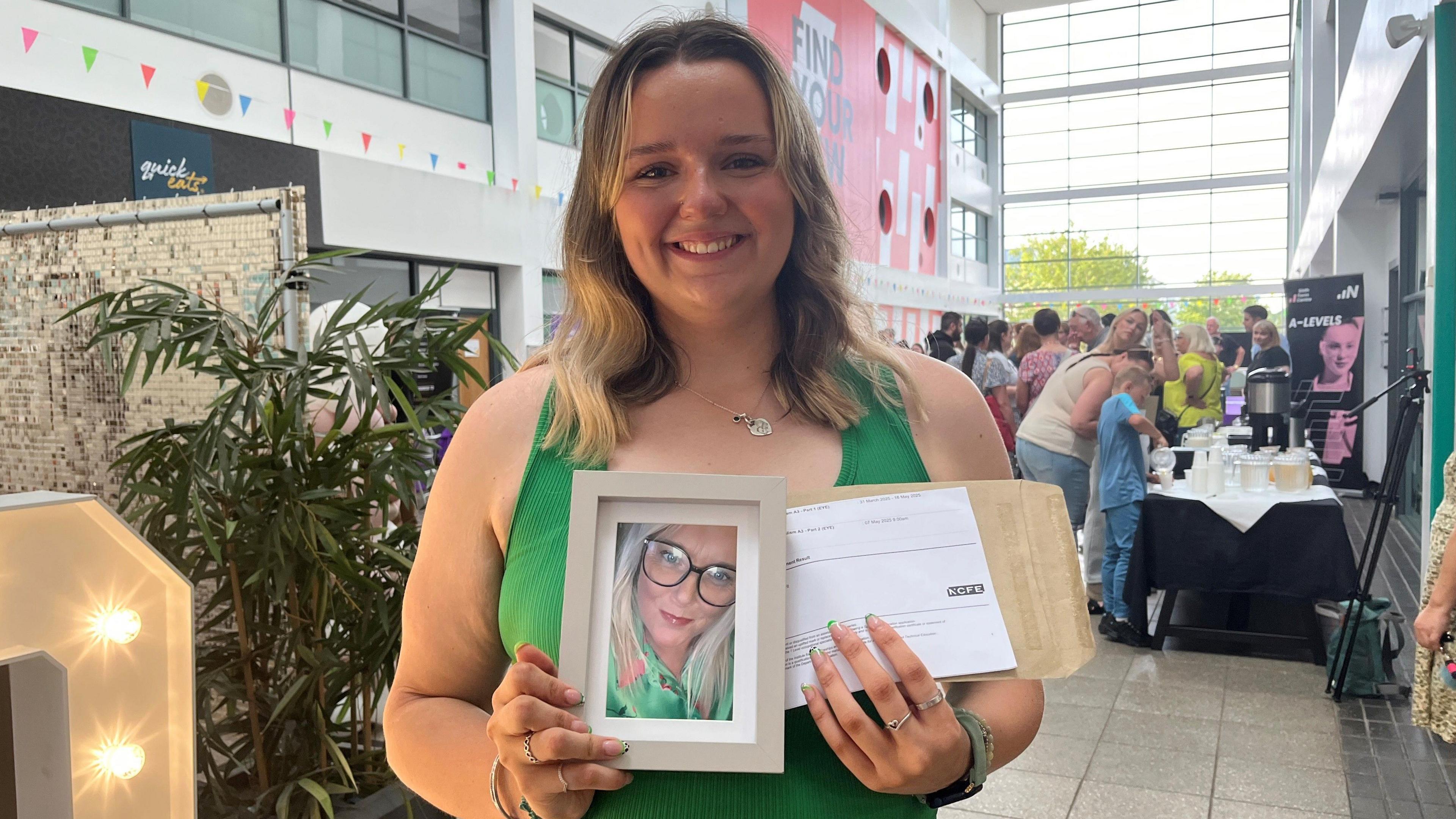 Molly Naas is smiling wearing a sleeveless green top. She is holding a picture of her late mother who is smiling and wearing glasses in the photo. She is also holding the paper with her exam results. There is a plant at the side and lots of people in the background at a coffee and tea stall. 