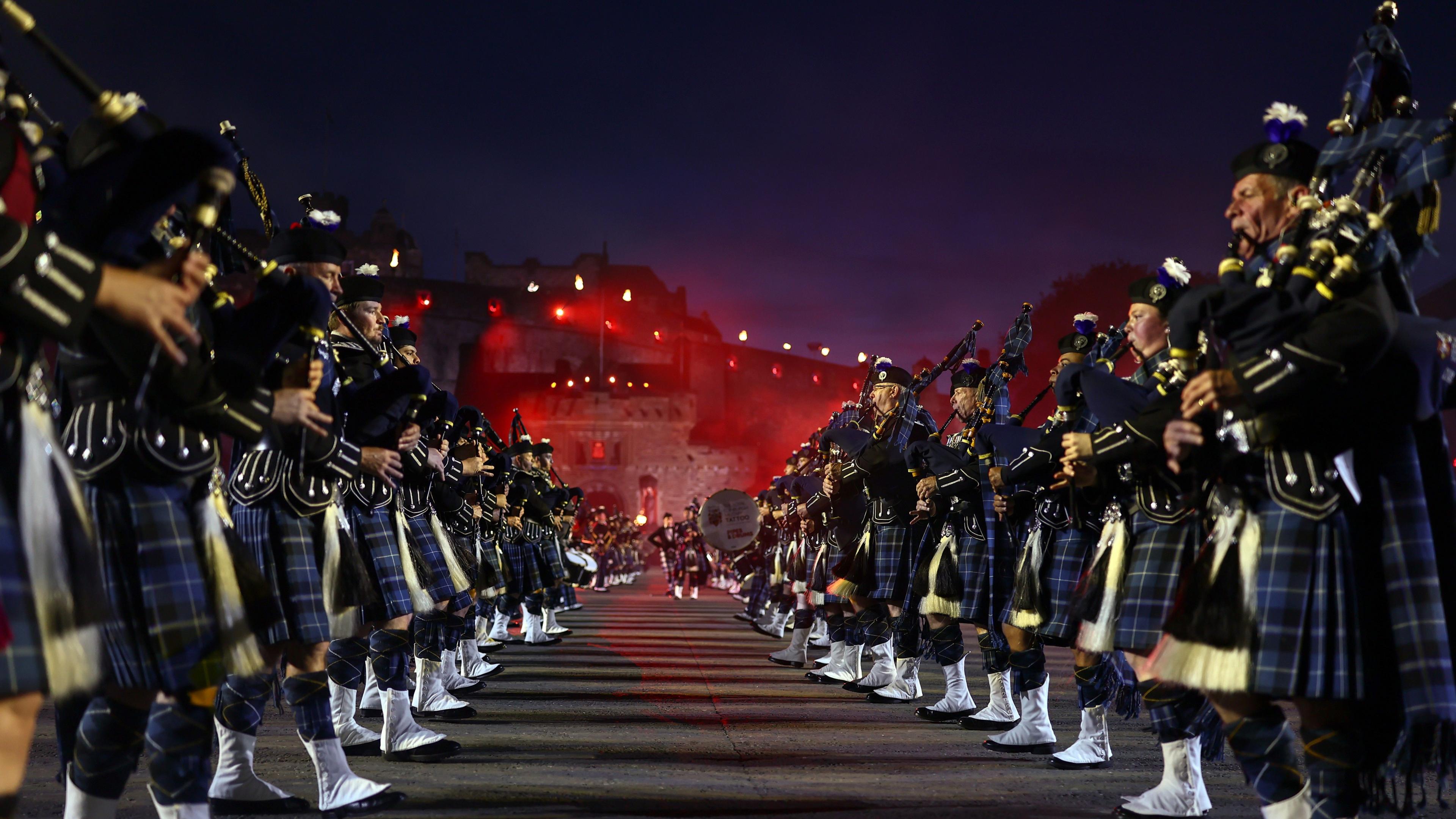 Two rows of pipers during a preview of the Royal Edinburgh Military Tattoo at Edinburgh Castle, which can be seen in the background shrouded in red light.