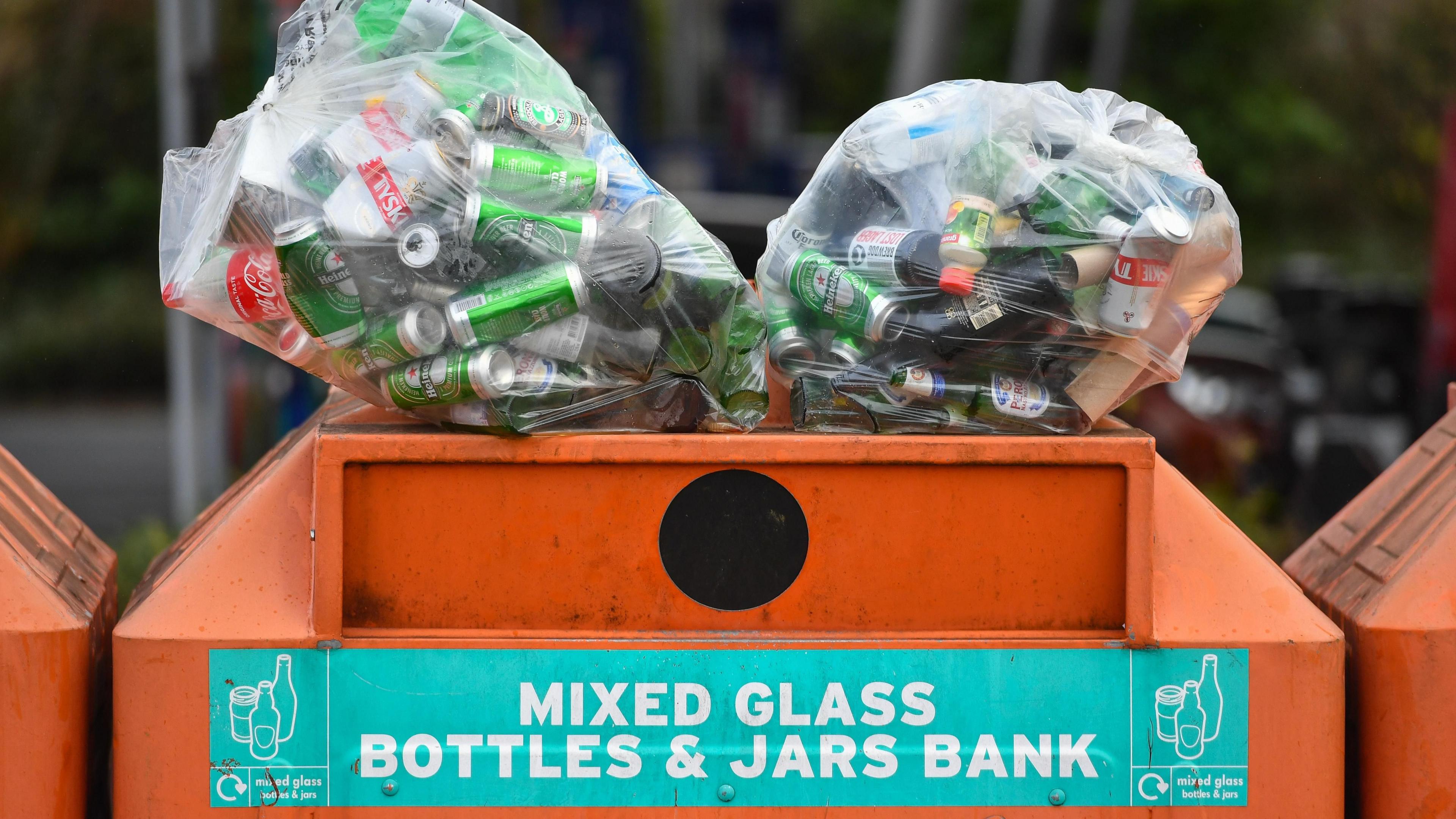 A large orange metal recycling bin that says "mixed glass bottles & jars bank" on it. On top of the bin are two clear plastic bags full of empty bottles and cans.