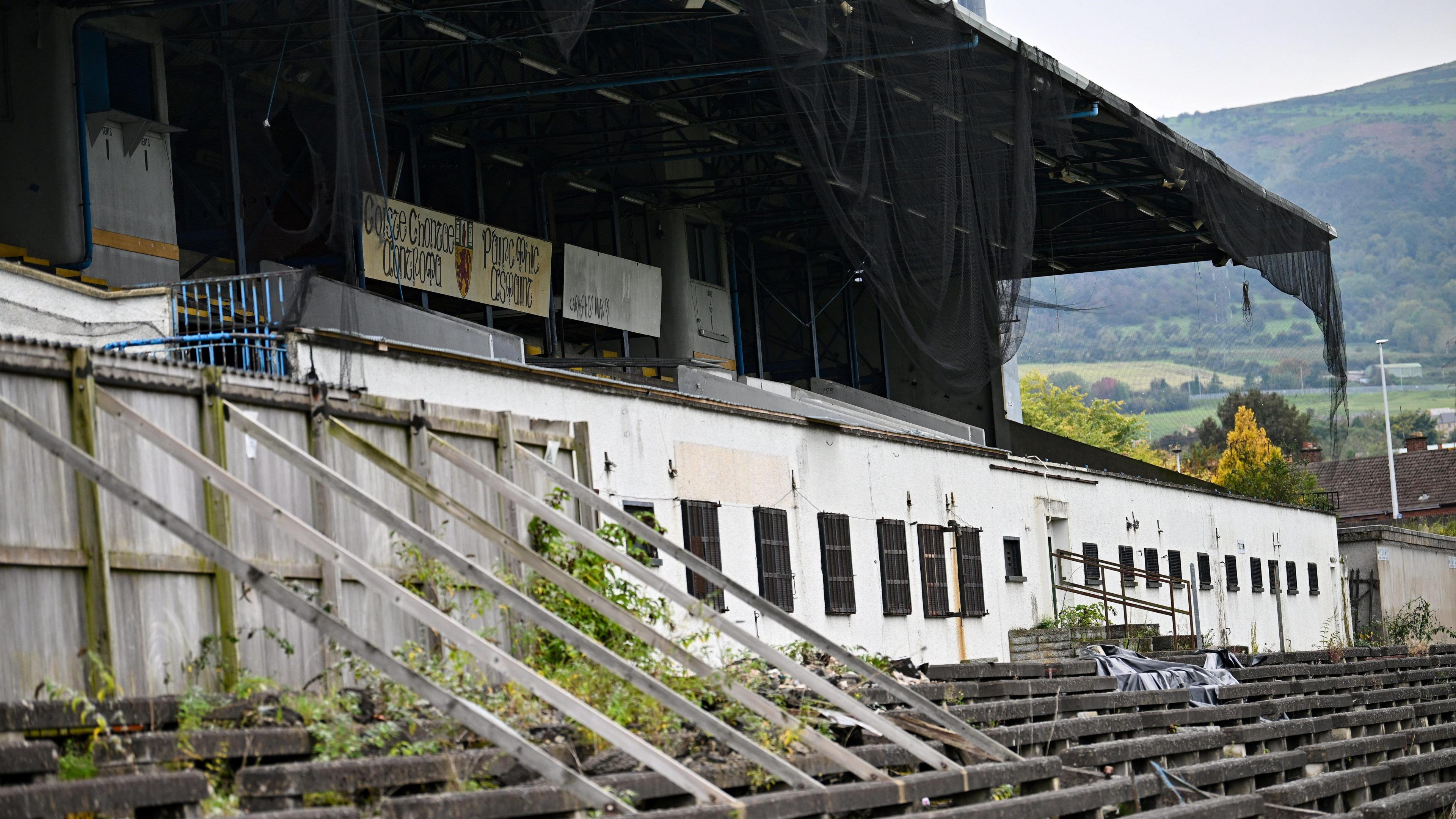A rundown stadium. Planks of wood are attached to a fence to keep it erect. Netting is hanging from the roof. Plants are overgrown.