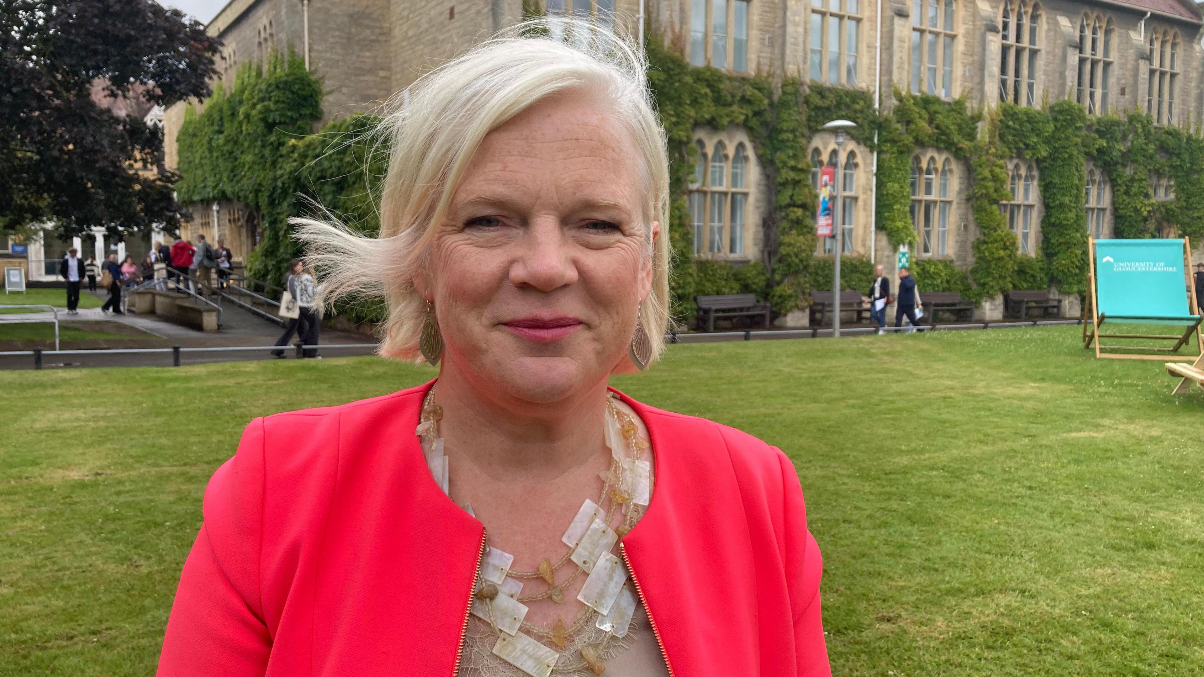 Clare Marchant stood on a historic University of Gloucestershire campus in Cheltenham. A vine-covered stone building stands behind her and students can be seen walking around. Ms Marchant is wearing a bright red jacket, a layered necklace, and her blonde hair is in a bob.