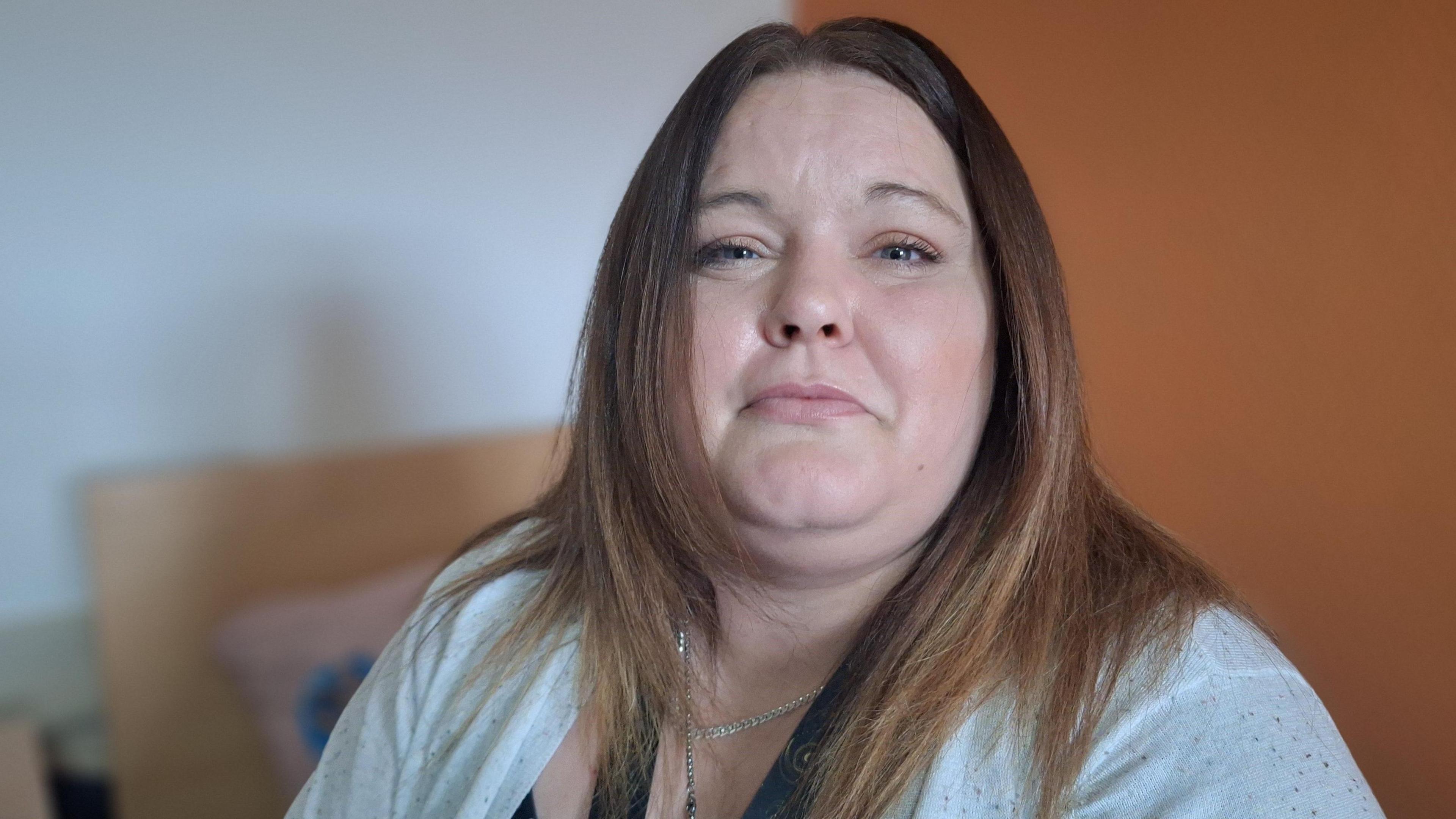 Charmaine Denton looking into the camera, smiling slightly. She has long straight brown/blond hair and is wearing a white cardigan and a silver chain. She is sat on the bed in her bedroom in the shared recovery focused residence she lives in.