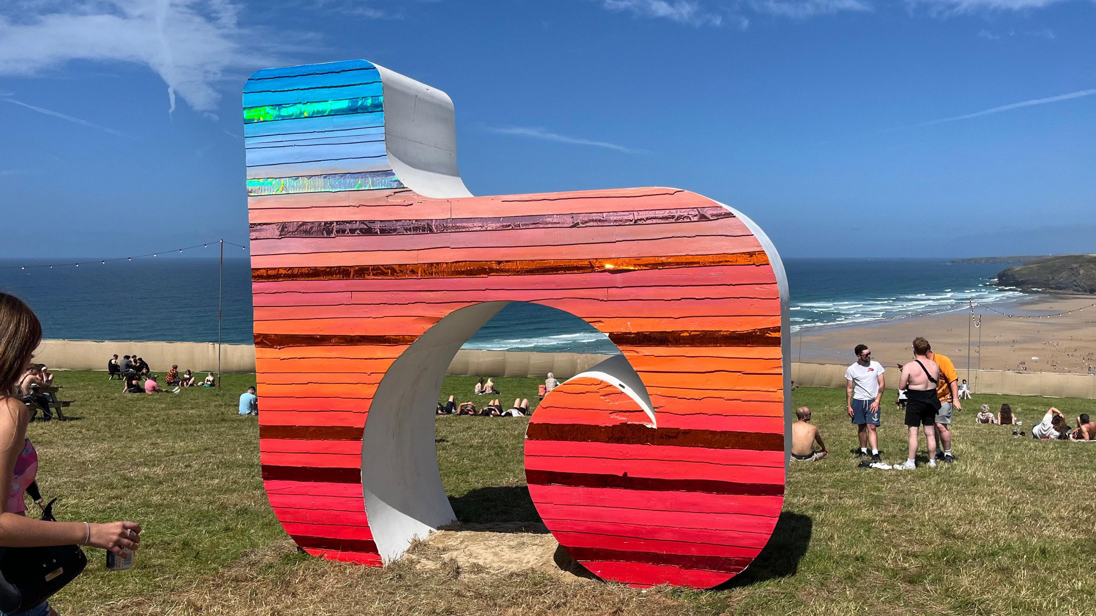 A blue, orange and red Boardmasters sign in a field overlooking a beach with people walking and sitting around it.