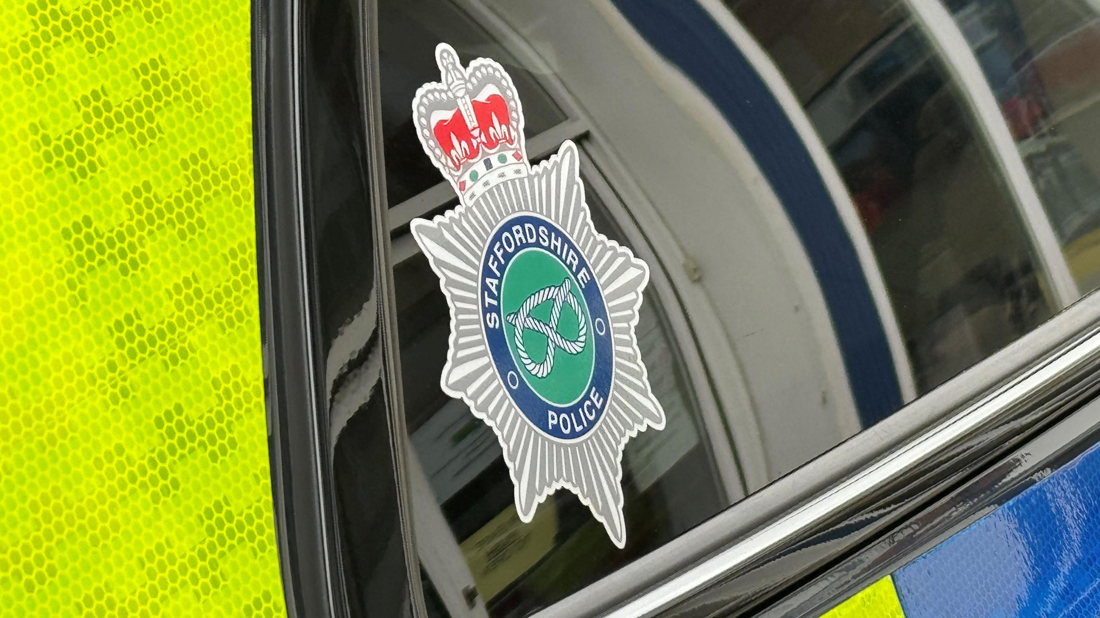 A Staffordshire Police crest on the rear window of a police car. The vehicle has yellow high visibility panels.